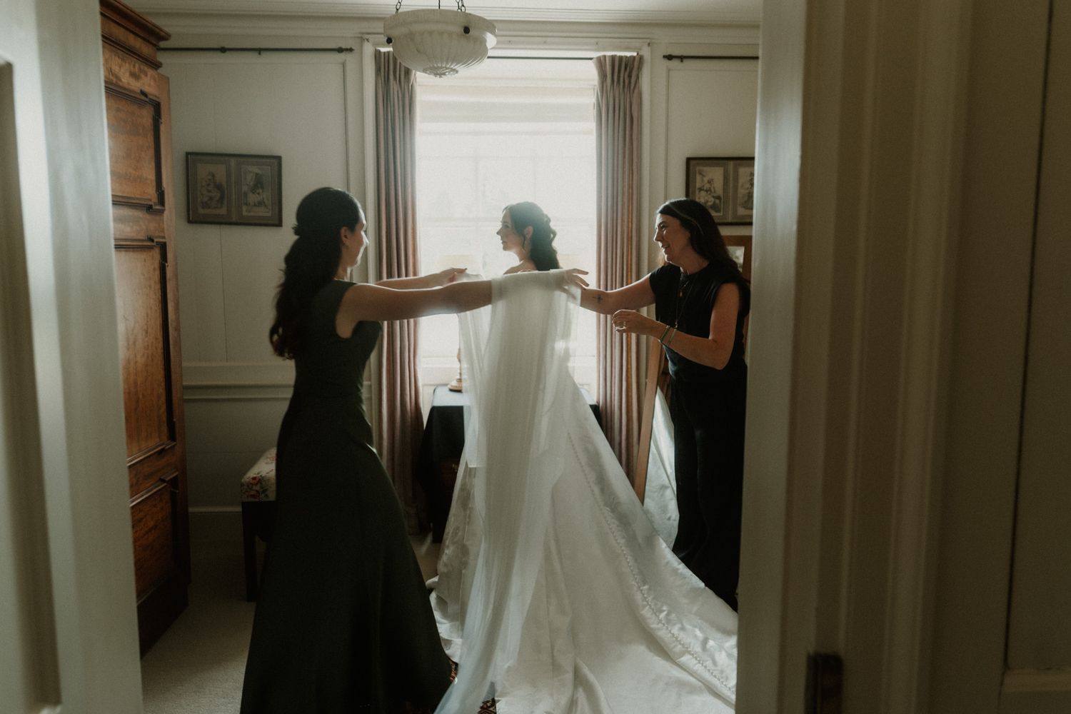 Bride in white gown being helped by two bridesmaids in black dresses near a bright window.