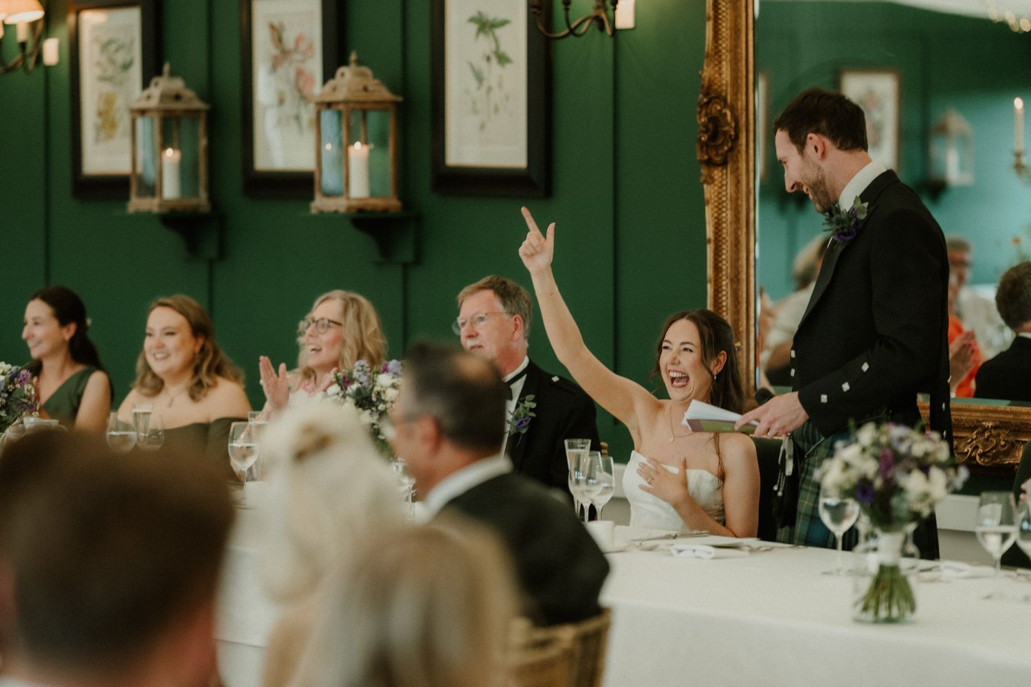 Wedding guests laughing and celebrating during a speech in an elegant green-walled reception room.
