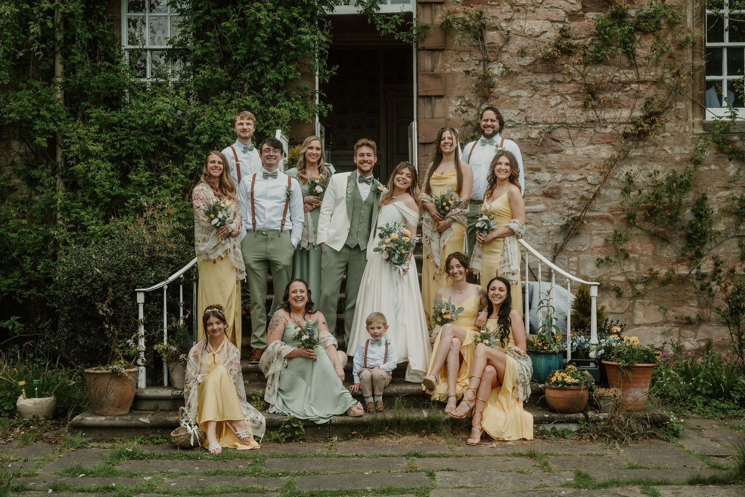 Wedding party poses on stone steps of ivy-covered venue, bride and groom centered among bridesmaids in yellow dresses.