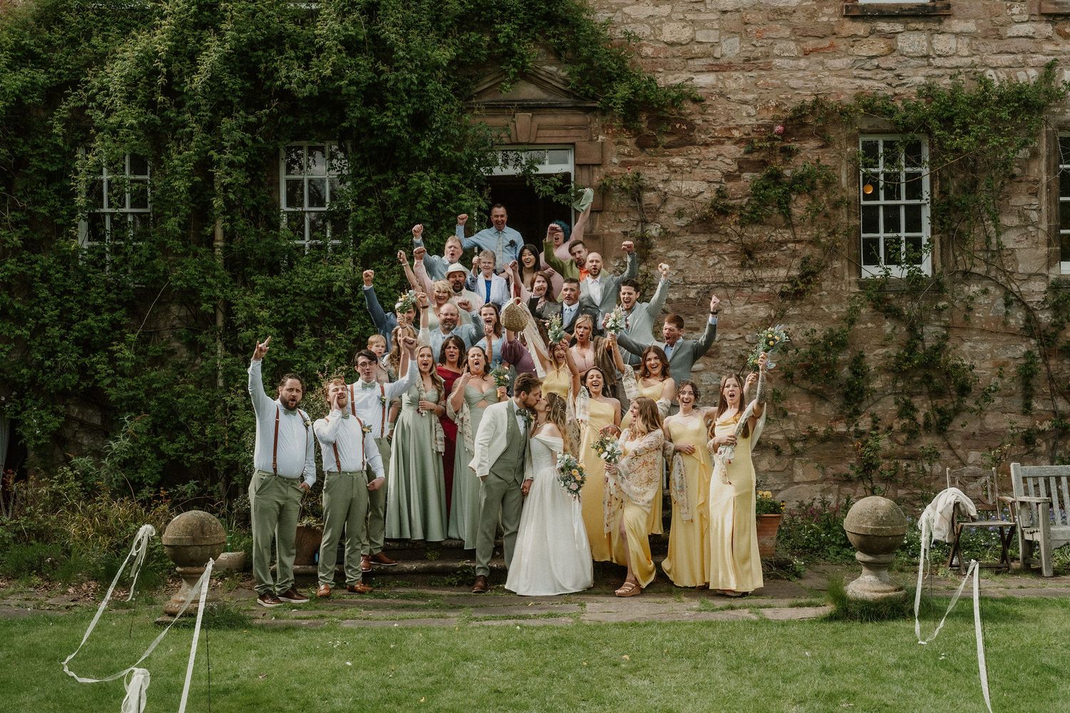 Wedding party celebrating on stone manor steps, bride in white, bridesmaids in yellow and sage green, arms raised joyfully.