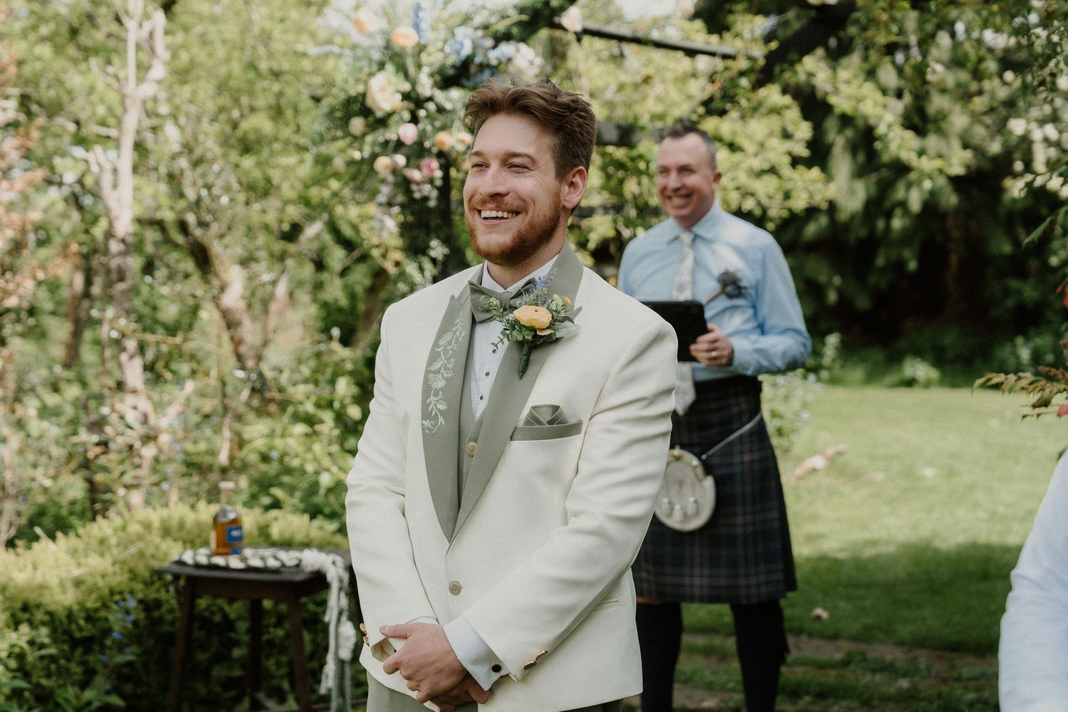 Smiling groom in ivory suit with floral boutonniere at outdoor wedding ceremony, man in kilt visible in background.