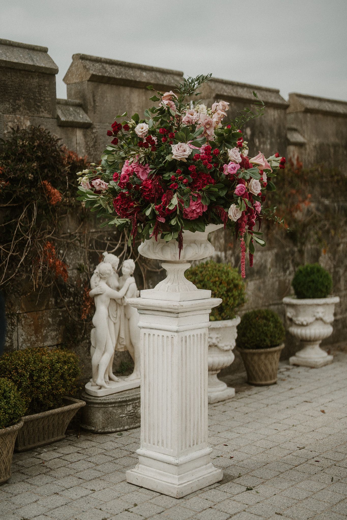 White classical columns and stone statues decorated with pink and red flowers along a stone pathway.