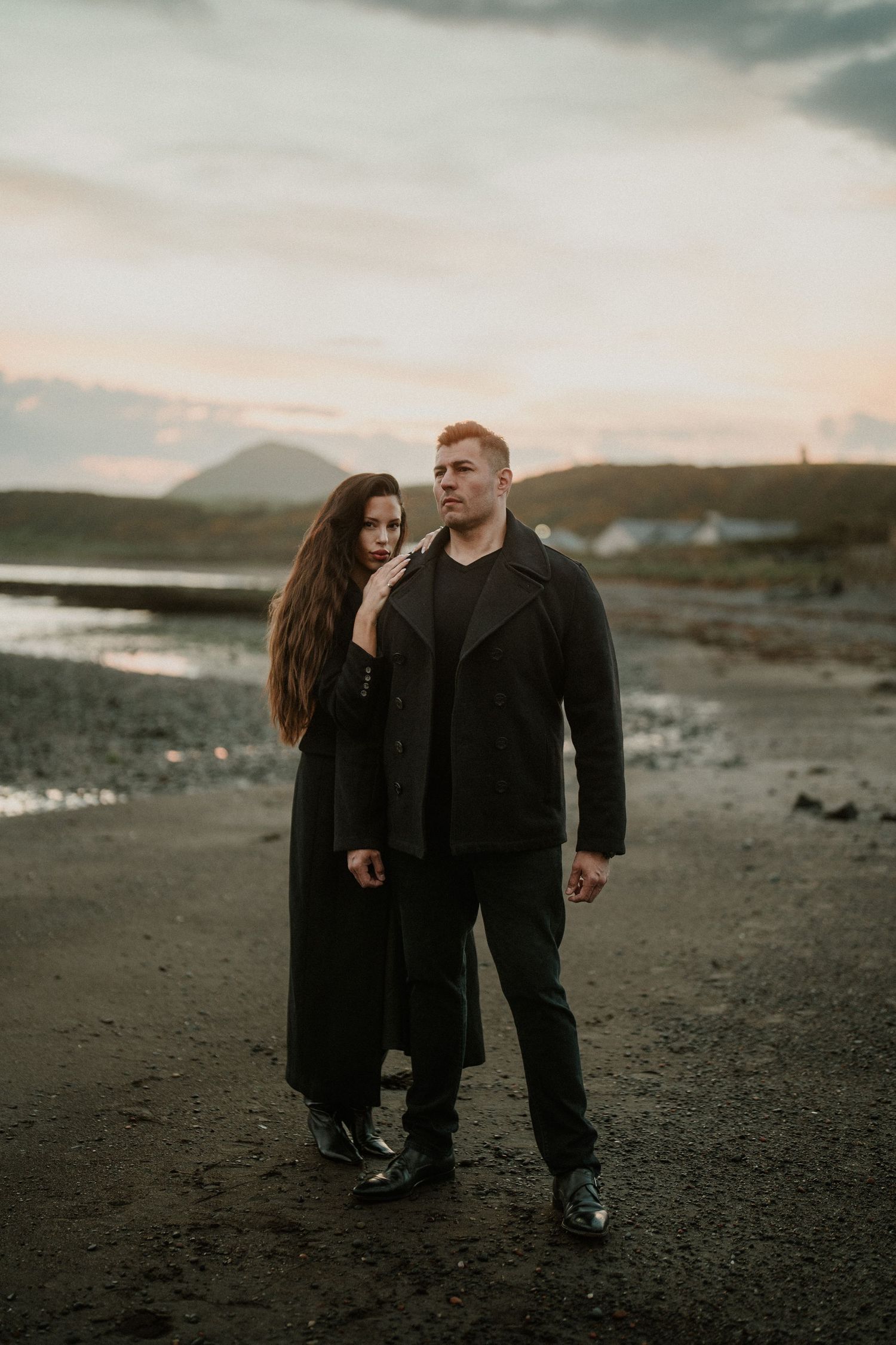 Couple in dark clothing poses on rocky beach at sunset with mountains in background.