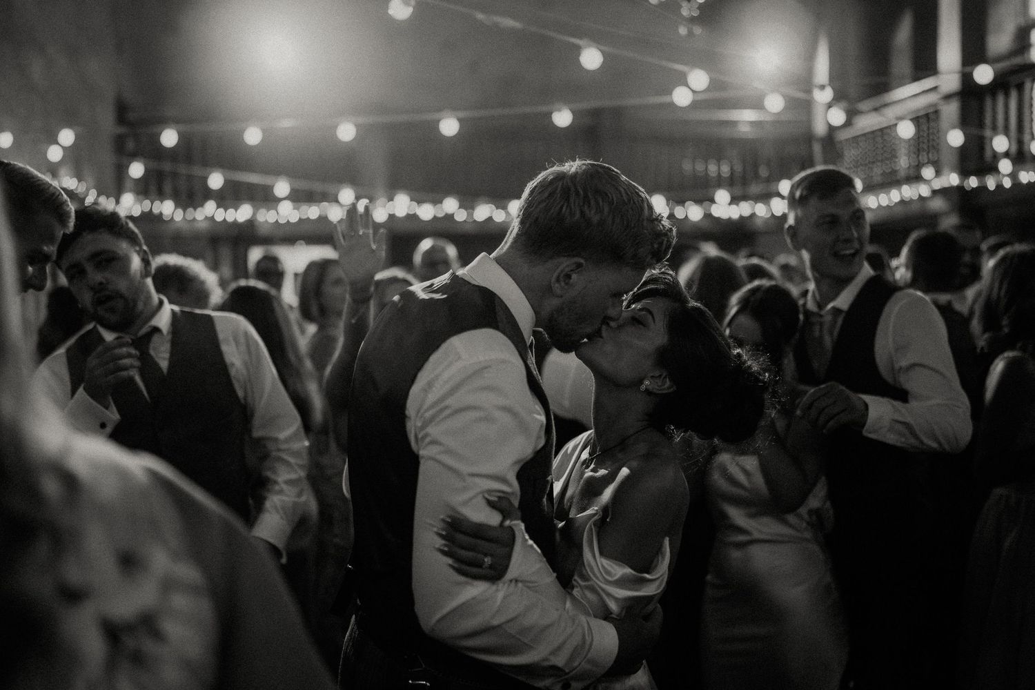 Silhouettes of wedding guests dancing under string lights at an evening reception.
