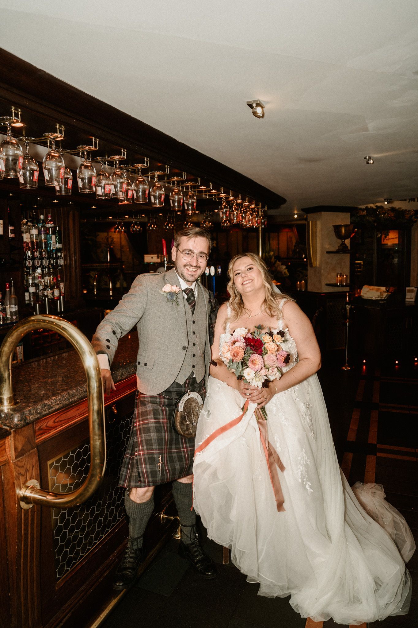 A wedding couple poses playfully in a dimly lit bar area with wood paneling and hanging lights.