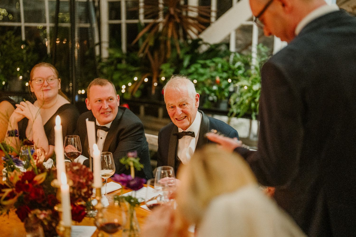 Guests share laughs and conversation around an elegantly decorated dinner table with candles and floral arrangements.