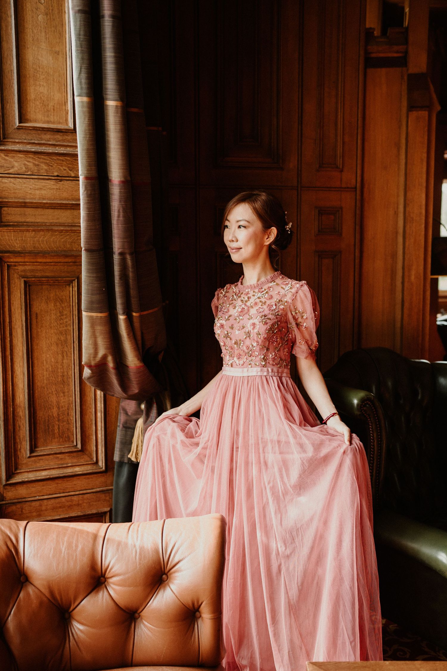 Woman in a pink vintage-style gown posing by wooden paneled walls and leather furniture.