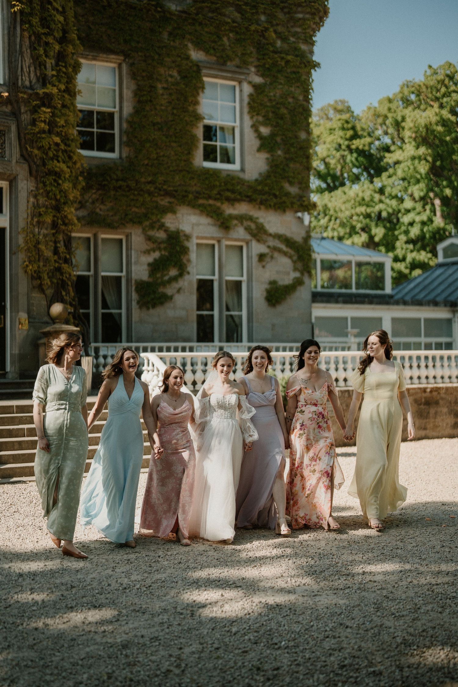 Wedding party in pastel dresses poses together on gravel path beside ivy-covered stone building.