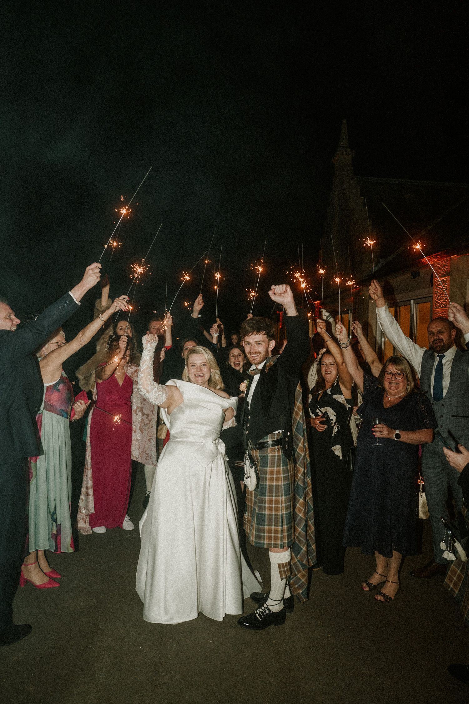 Wedding guests hold sparklers around a couple in traditional Scottish and white wedding attire during nighttime celebration.