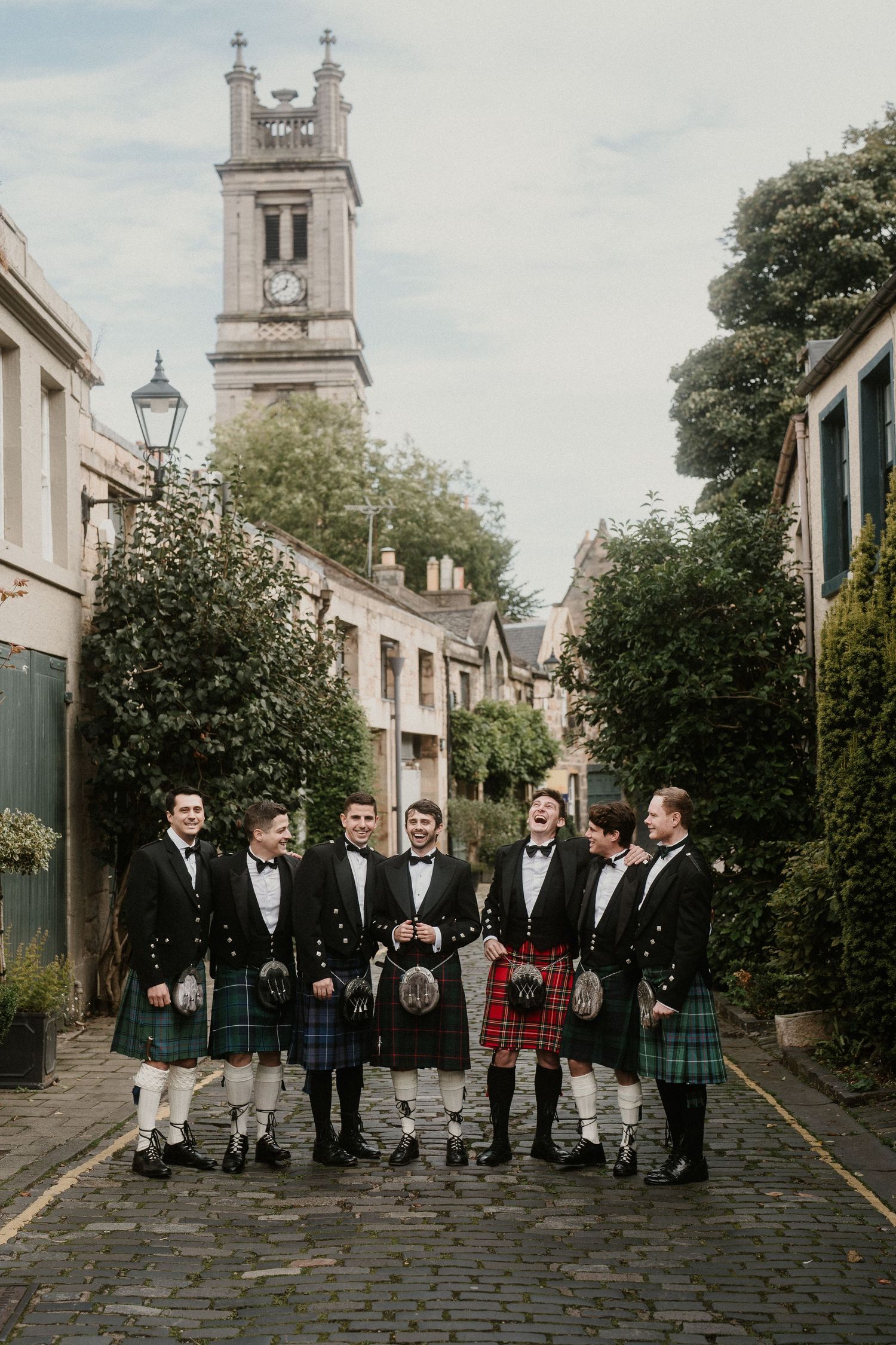 Wedding party wearing traditional Scottish kilts and formal attire pose on a cobblestone street with church tower backdrop.