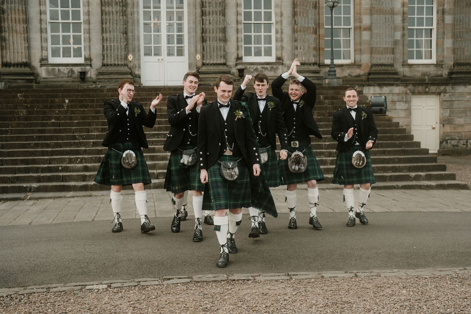 Group wearing kilts and formal attire walk in formation on stone courtyard of grand estate.