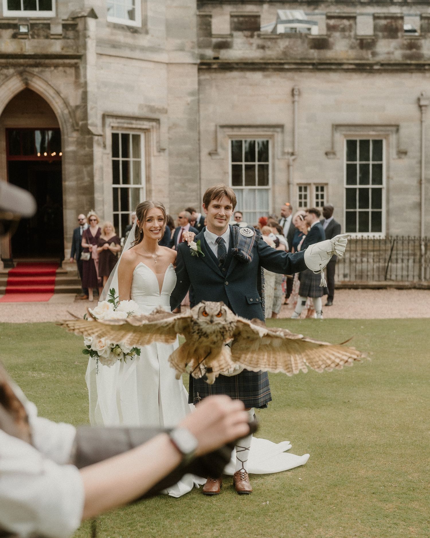 Bride and groom pose with a large owl in flight at a Scottish castle wedding, groom wearing traditional kilt.