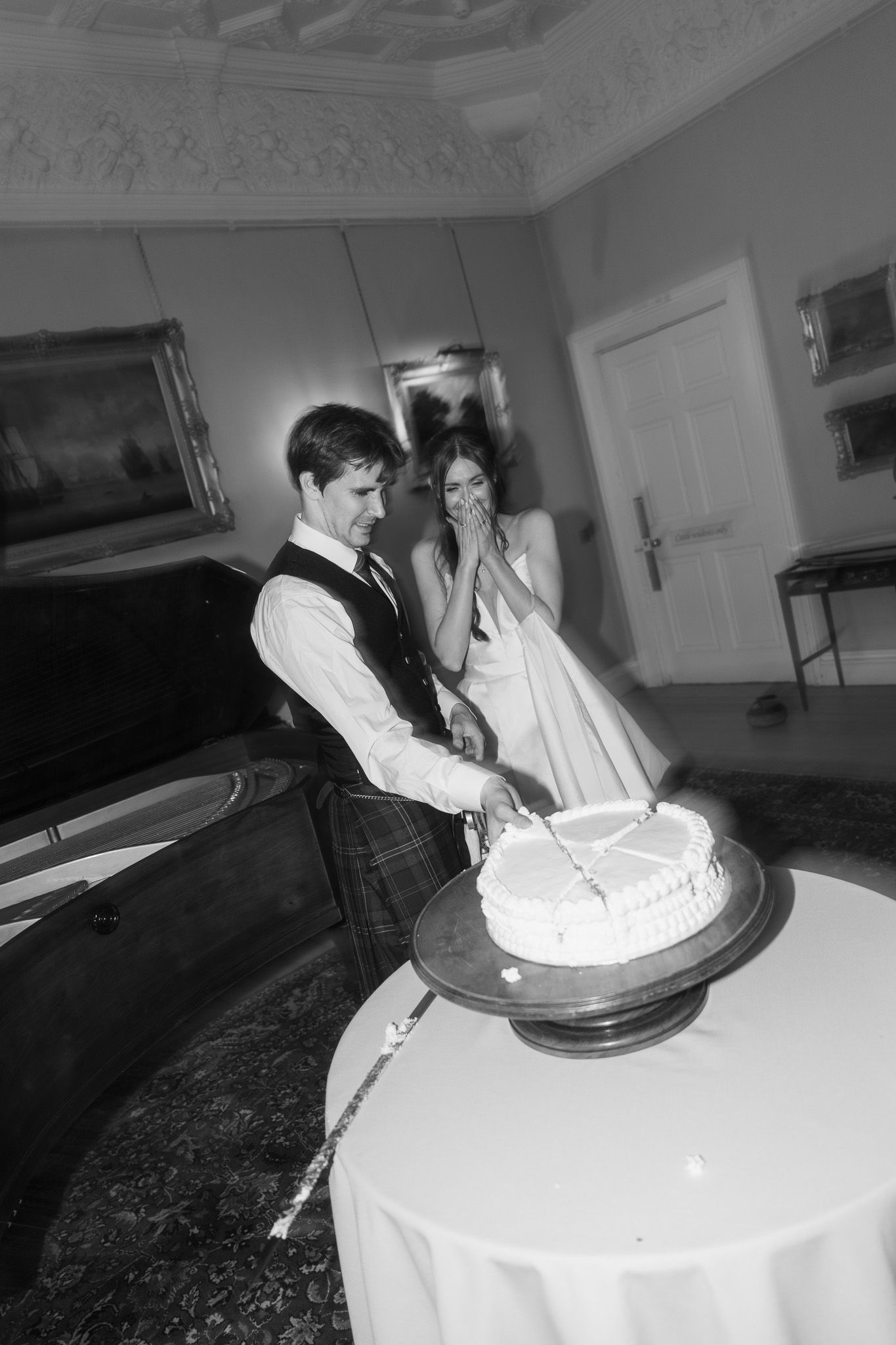 Black and white photo of bride and groom cutting wedding cake in elegant venue, groom wearing tartan kilt.