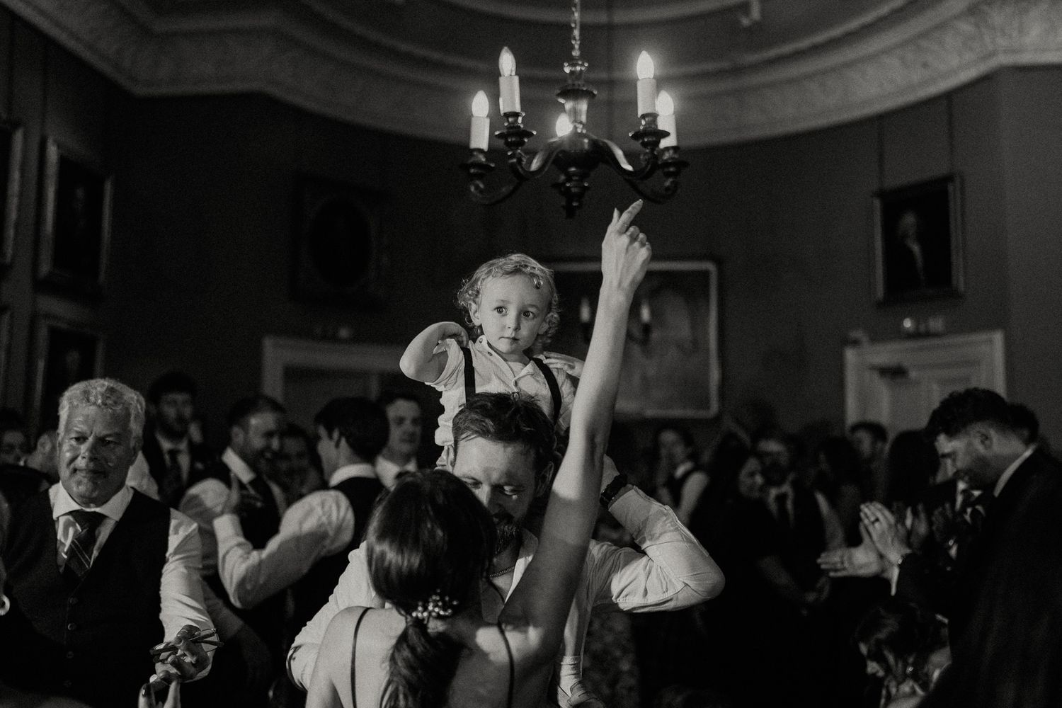 Black and white wedding reception photo of child on shoulders near chandelier amid dancing guests.
