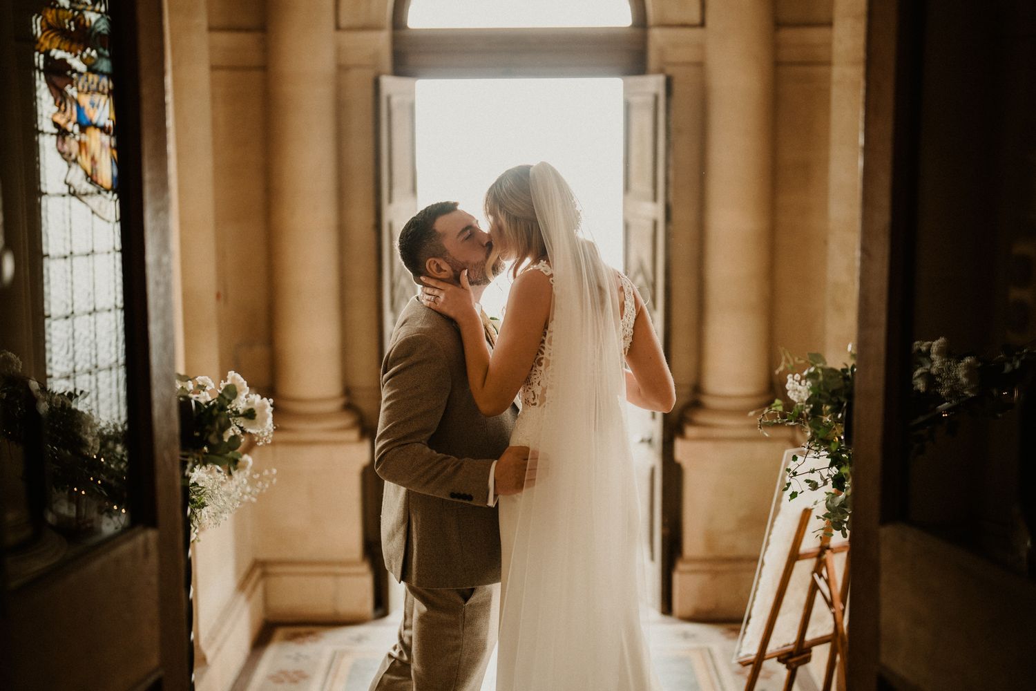 A bride and groom share a romantic kiss in a sunlit doorway, with the bride in a white gown and veil.