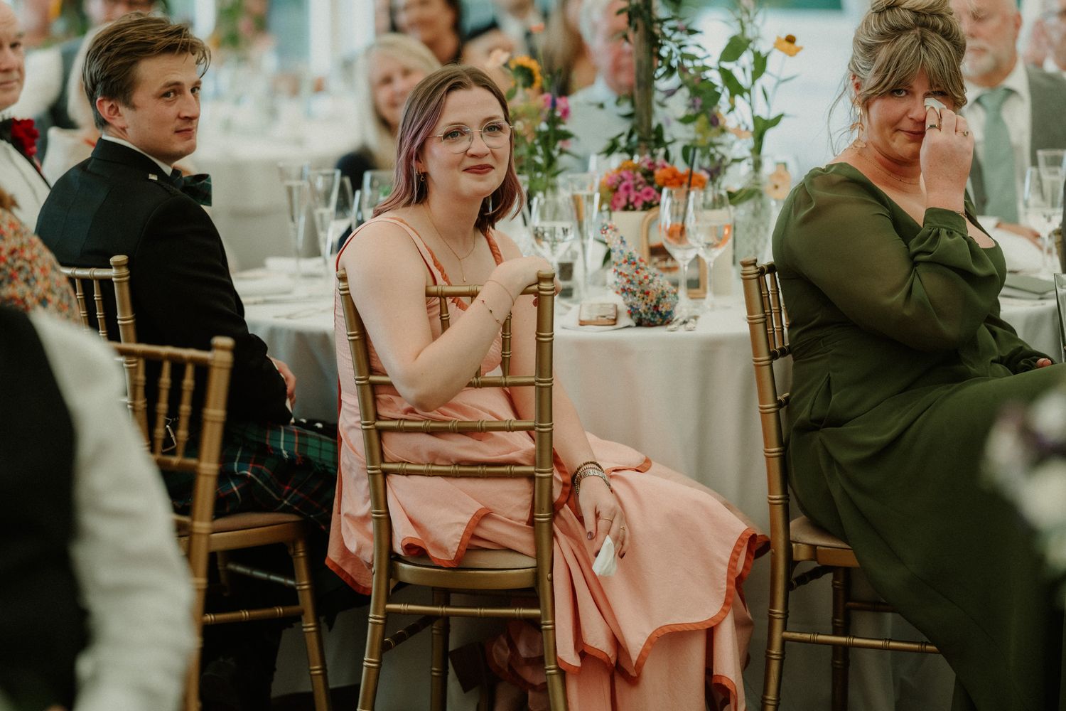 Guests seated at wedding reception tables, woman in pink dress laughing, another in green dress emotional.
