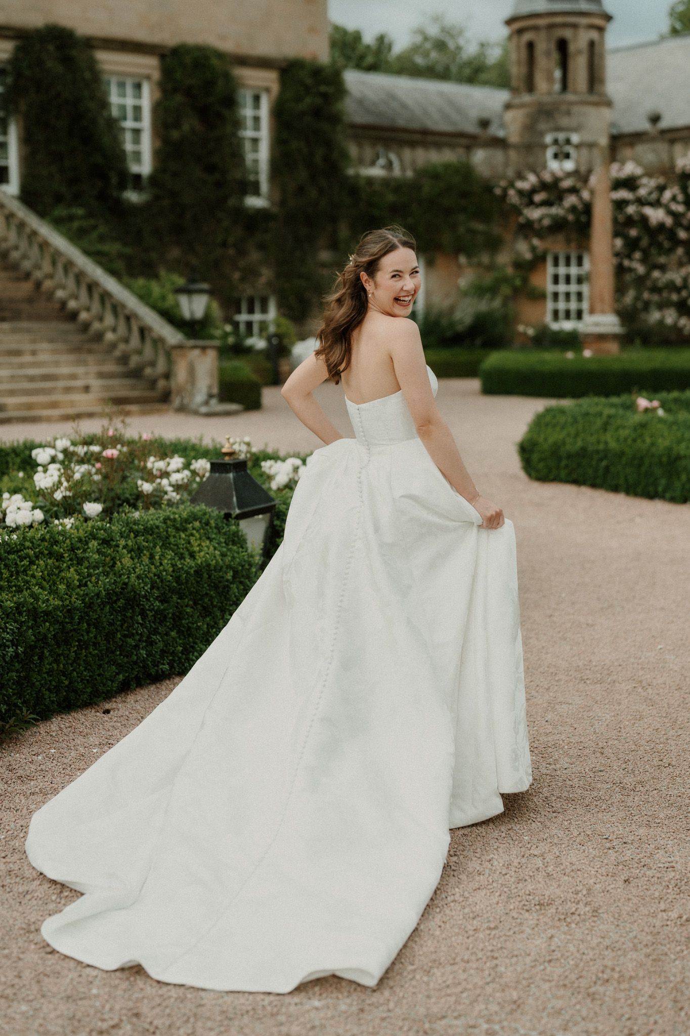Bride in strapless white ballgown twirling in garden of elegant stone manor house.