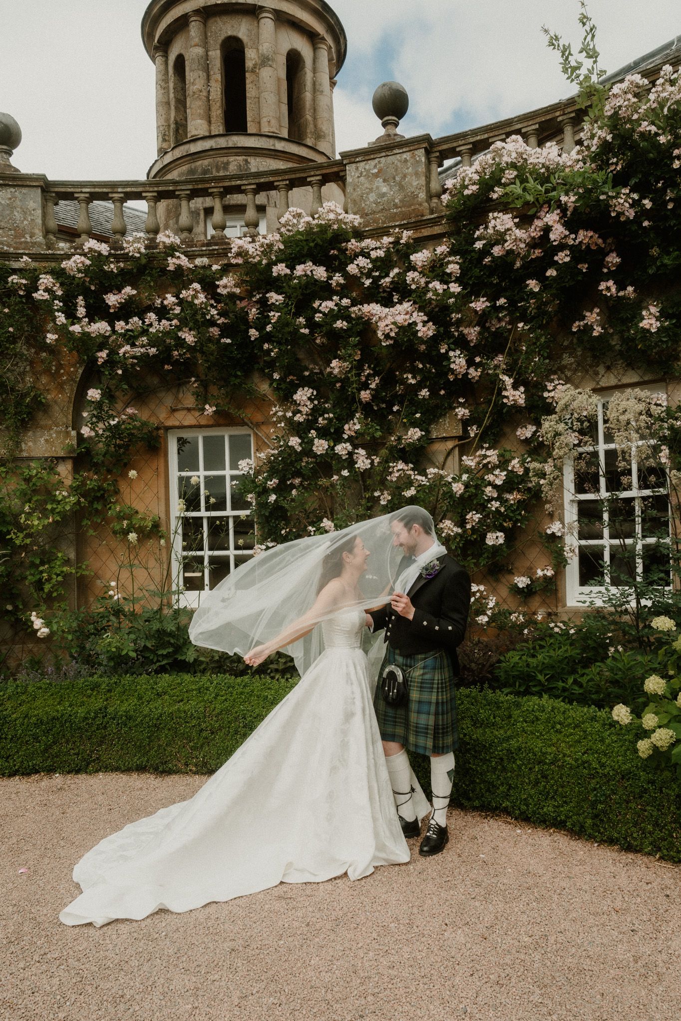 Bride in white gown and groom in Scottish kilt share a kiss under veil at a floral-covered stone manor.
