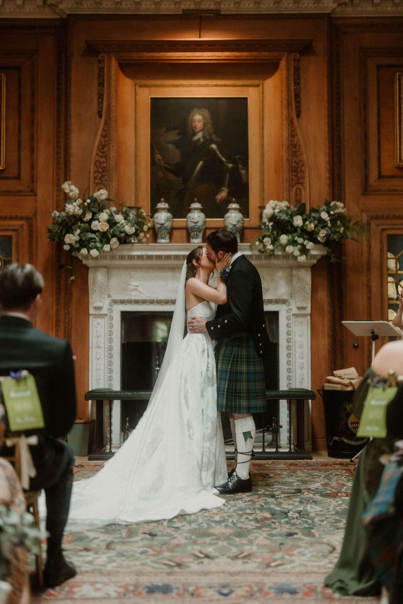Bride and groom share first kiss at elegant Scottish castle wedding ceremony with floral arrangements and portrait above fireplace.