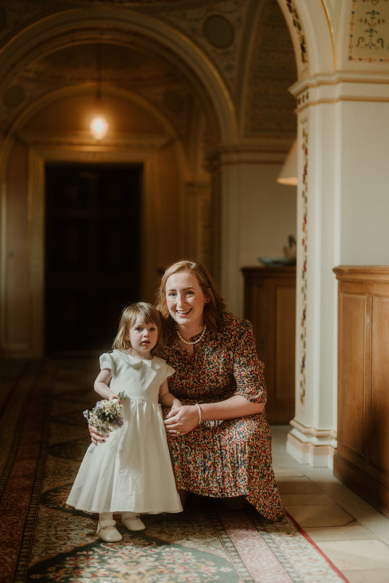 Woman in floral dress crouches beside young flower girl in white dress inside an elegant church corridor.