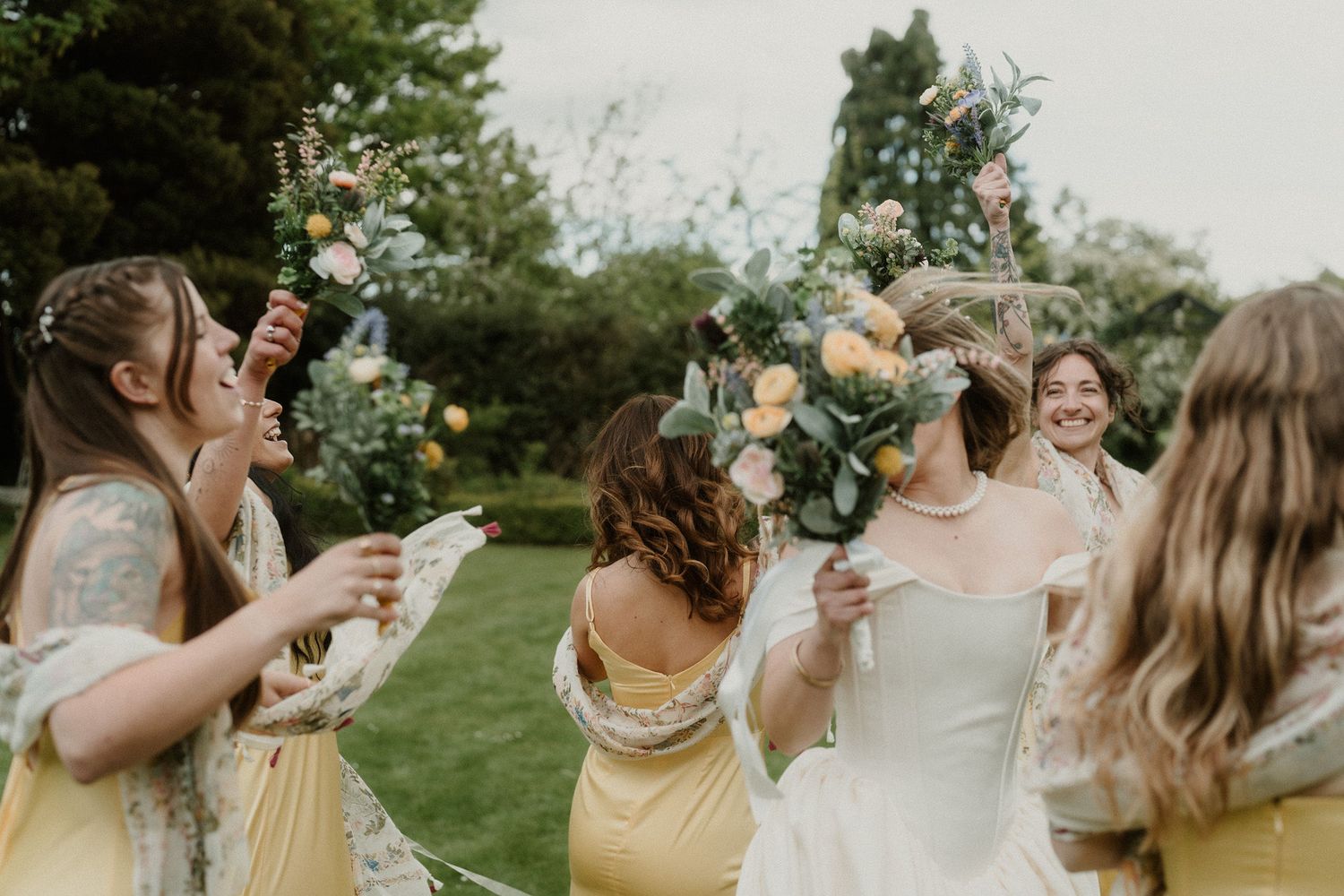 Bride in white gown celebrates with bridesmaids in yellow dresses, holding floral bouquets outdoors at a wedding.
