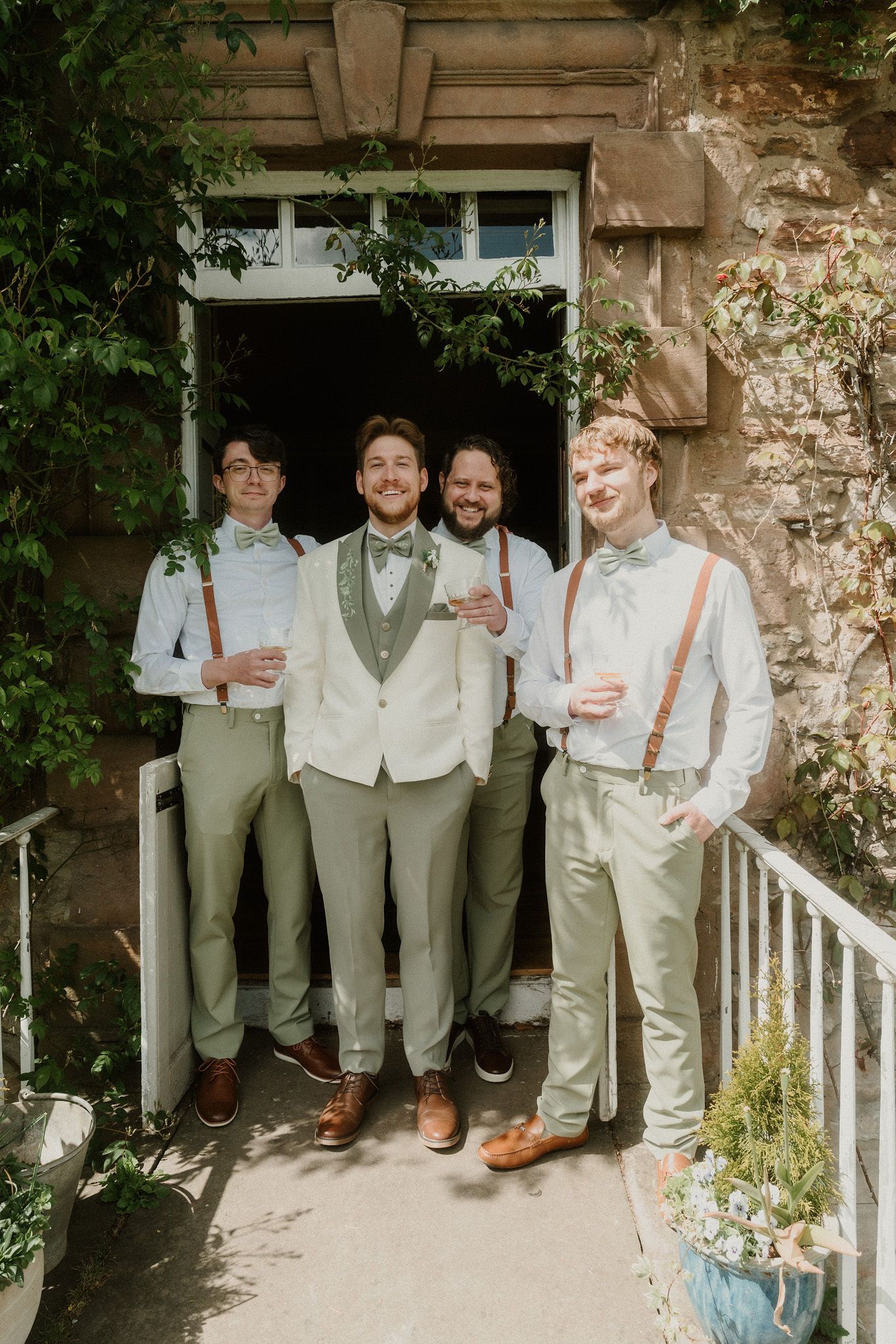 Groom and groomsmen in sage green suits with bow ties and suspenders pose outside a stone building.