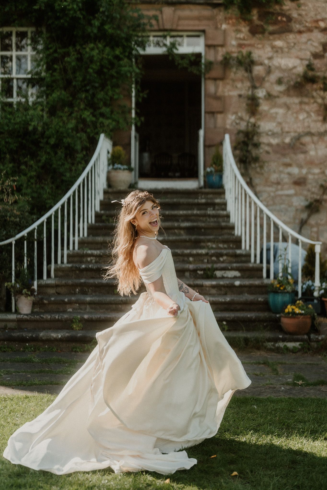 A bride in a flowing ivory off-shoulder gown twirls gracefully on a lawn before a stone manor with white railings.