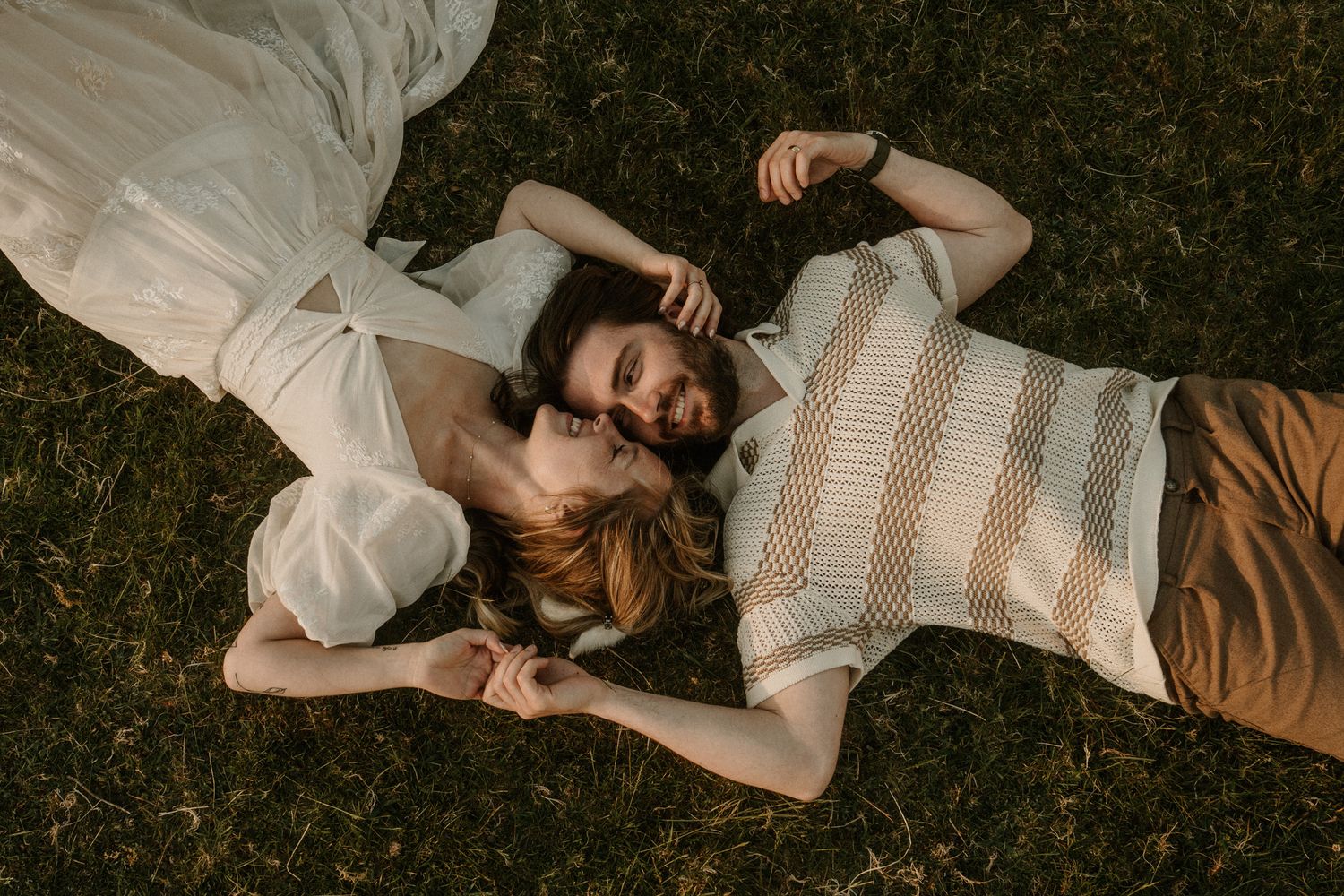 A couple lying on green grass, heads together, holding hands in a romantic outdoor setting.