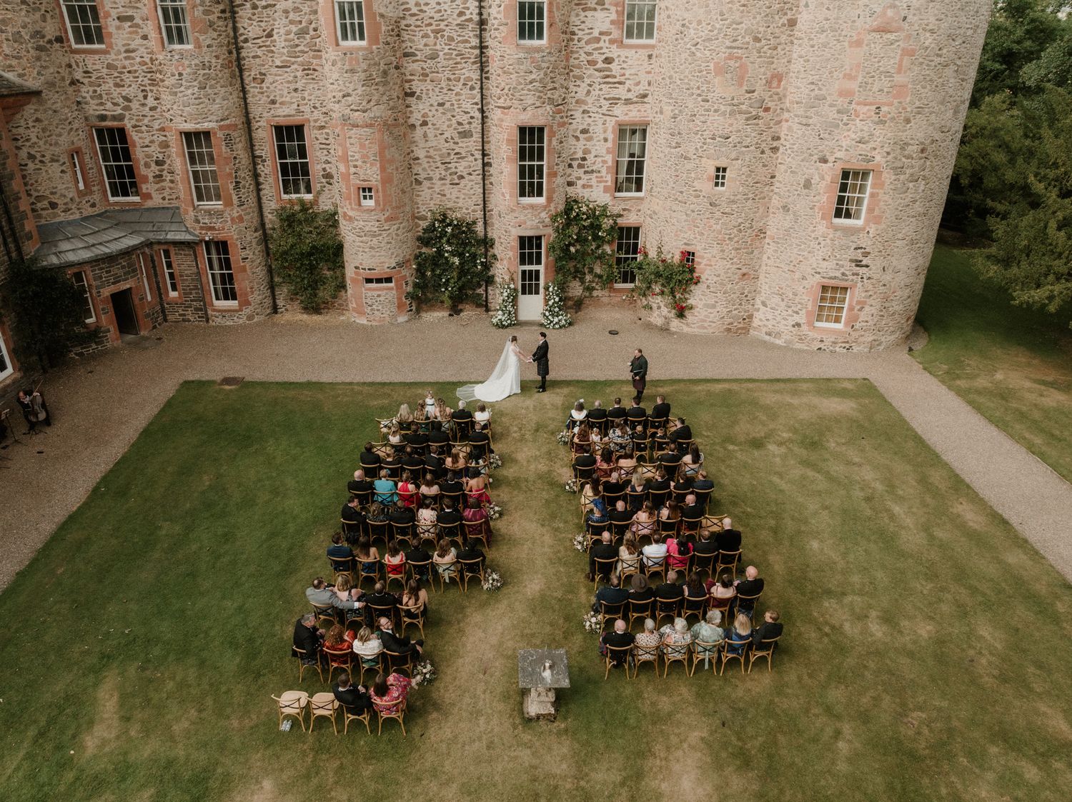 Aerial view of an outdoor wedding ceremony on a castle lawn with guests seated in two sections as the couple stands at the front.