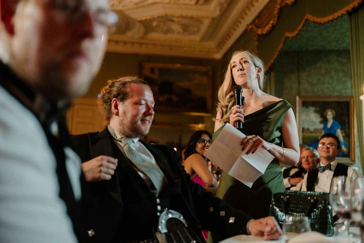 Woman in a green dress holds a microphone and paper while giving a speech to seated guests in an ornate, elegant banquet room.