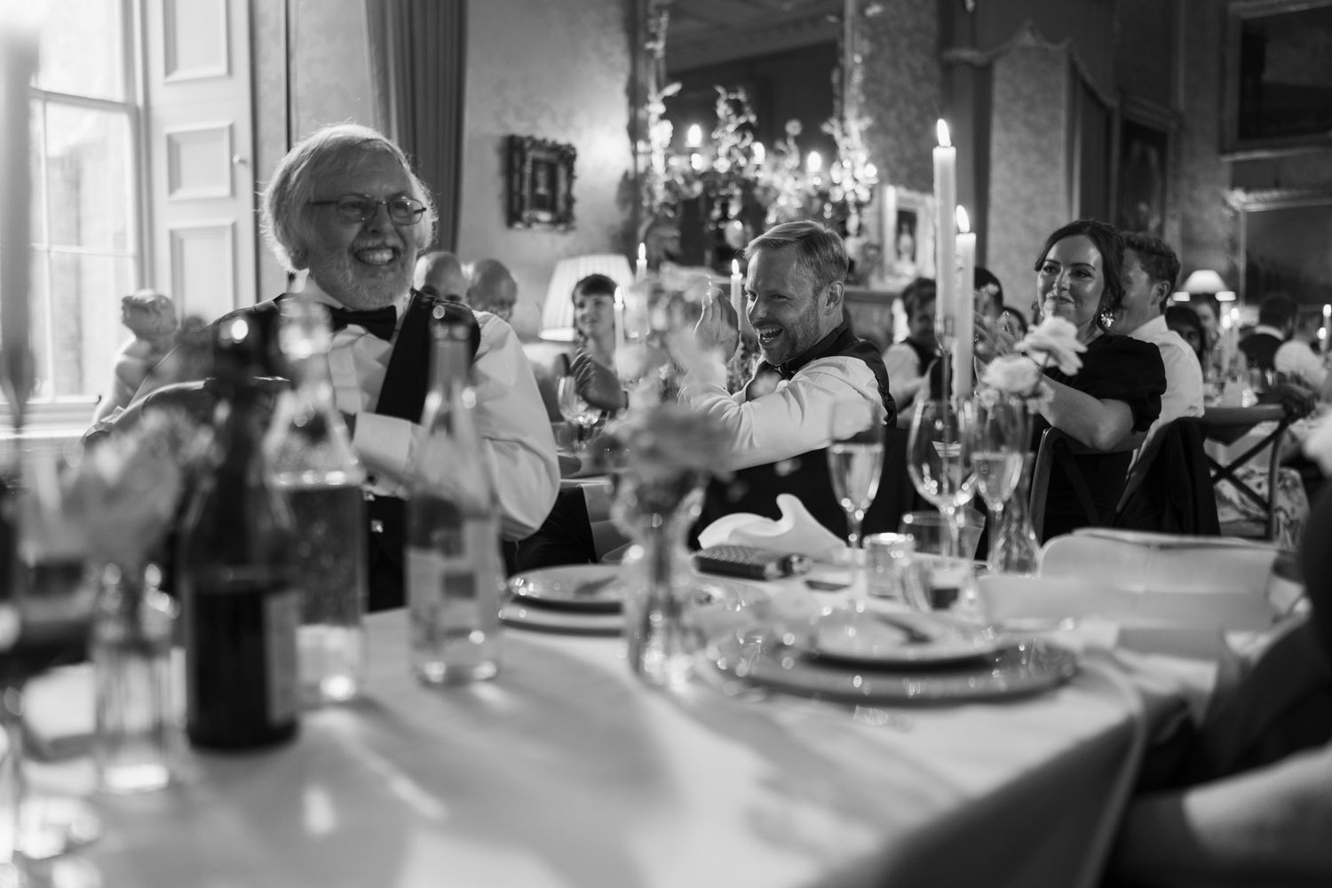 Black and white photo of guests laughing and applauding at a candlelit banquet table set with plates, wine glasses and bottles.