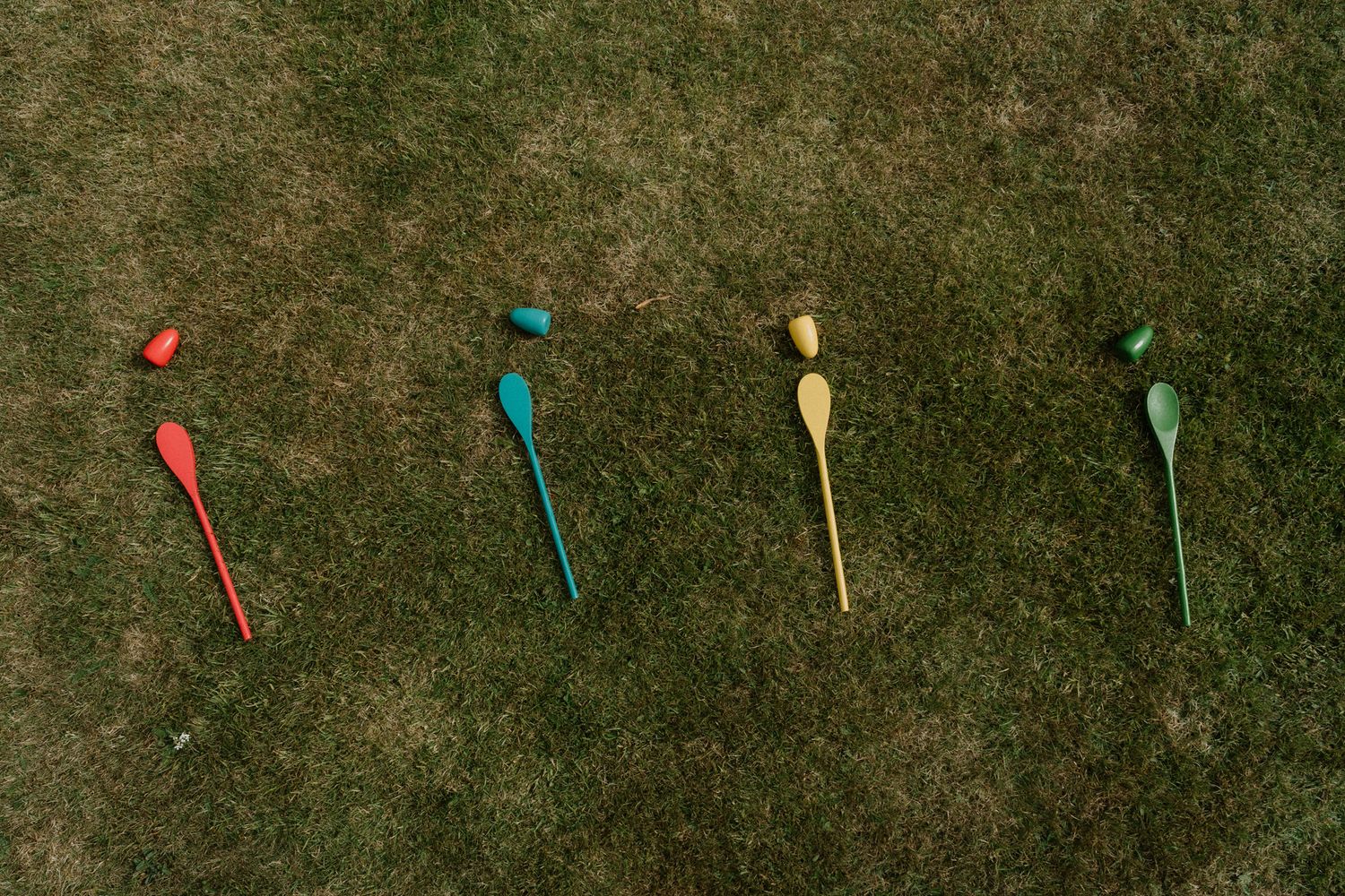 Four colorful wooden spoons in red, blue, yellow and green lie in a row on grass, each with a small matching egg or ball above its bowl.