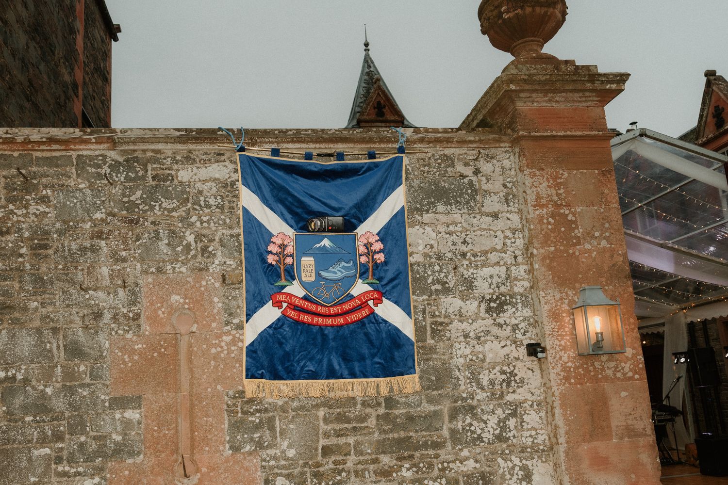 Blue heraldic banner with crest, white diagonal cross and gold fringe hanging on a weathered stone wall beside a wall-mounted lantern.