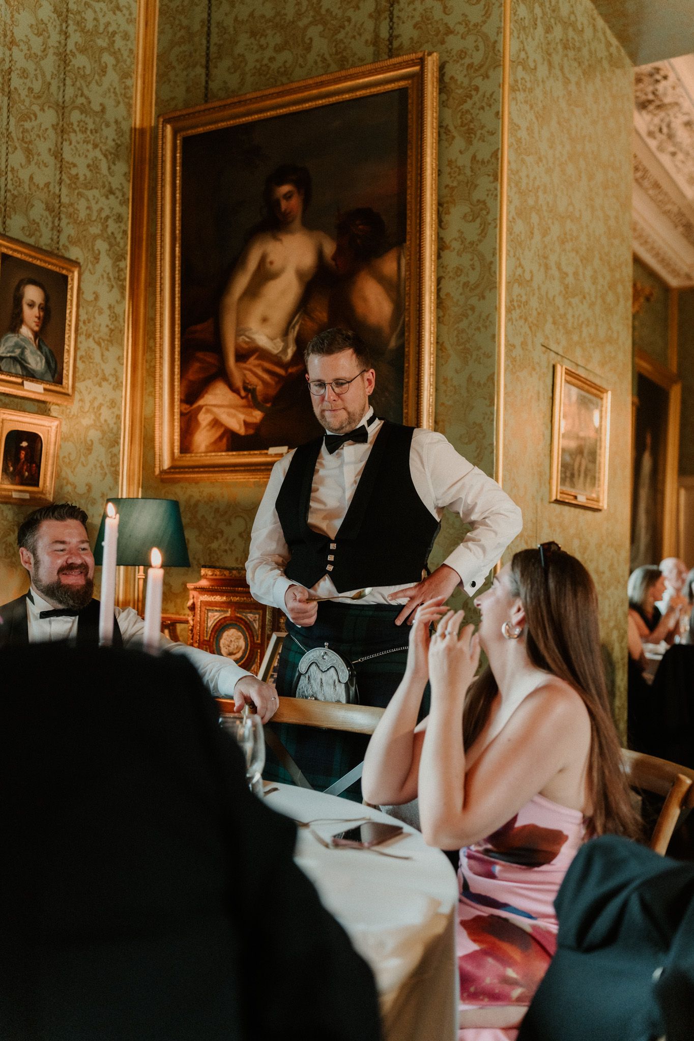 Waiter in black vest and bow tie stands beside a seated woman in a pink dress at a candlelit table in an ornate room with gilded paintings.