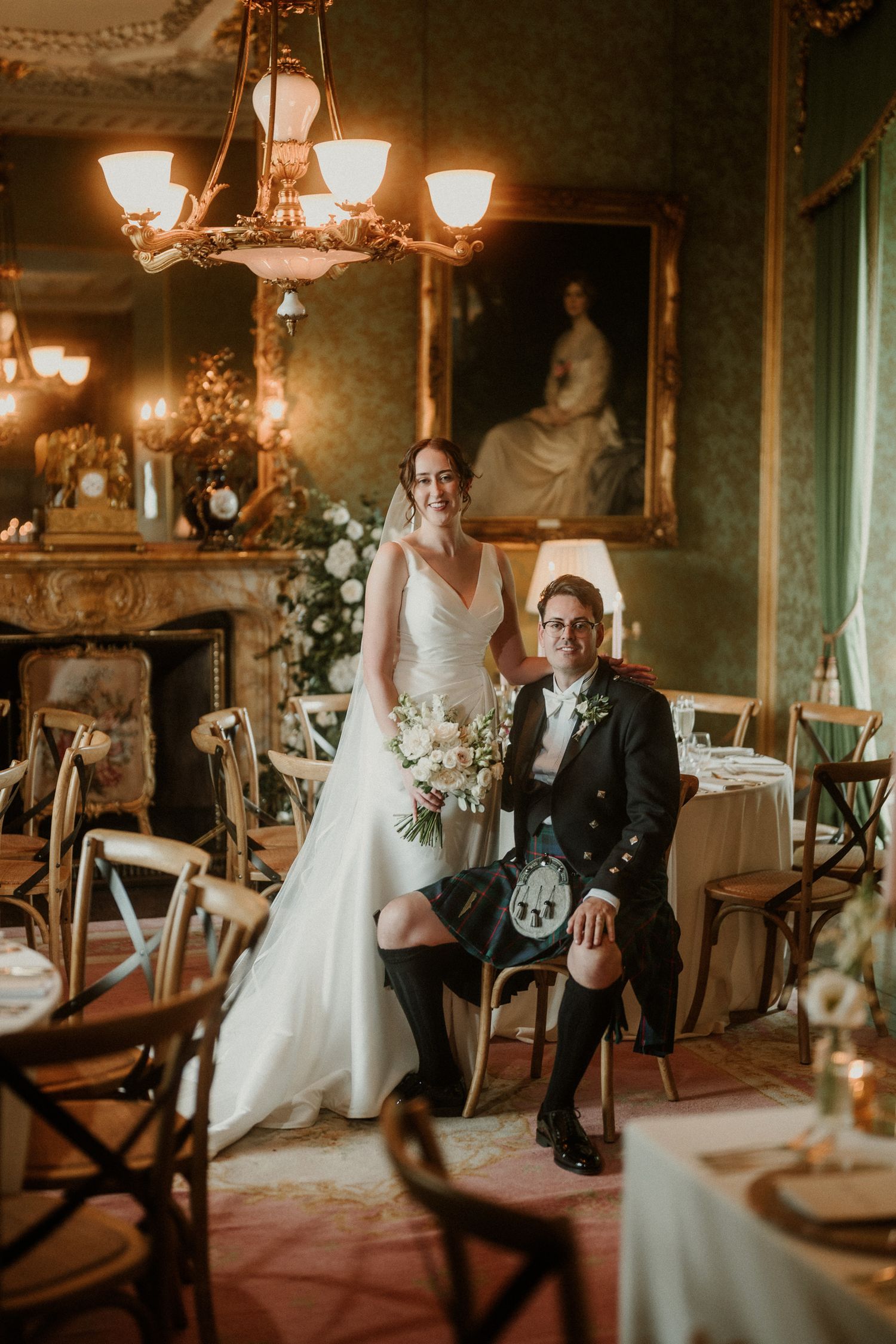 Bride and groom pose in elegant Scottish castle wedding venue, groom wearing traditional kilt, bride holding white bouquet.