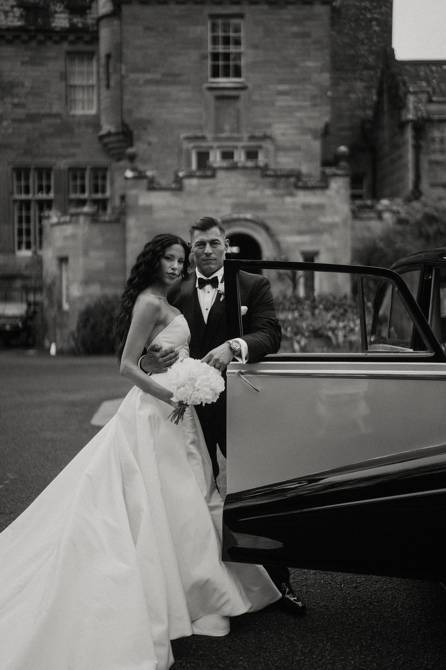 Elegant couple poses with vintage black car outside historic stone building.