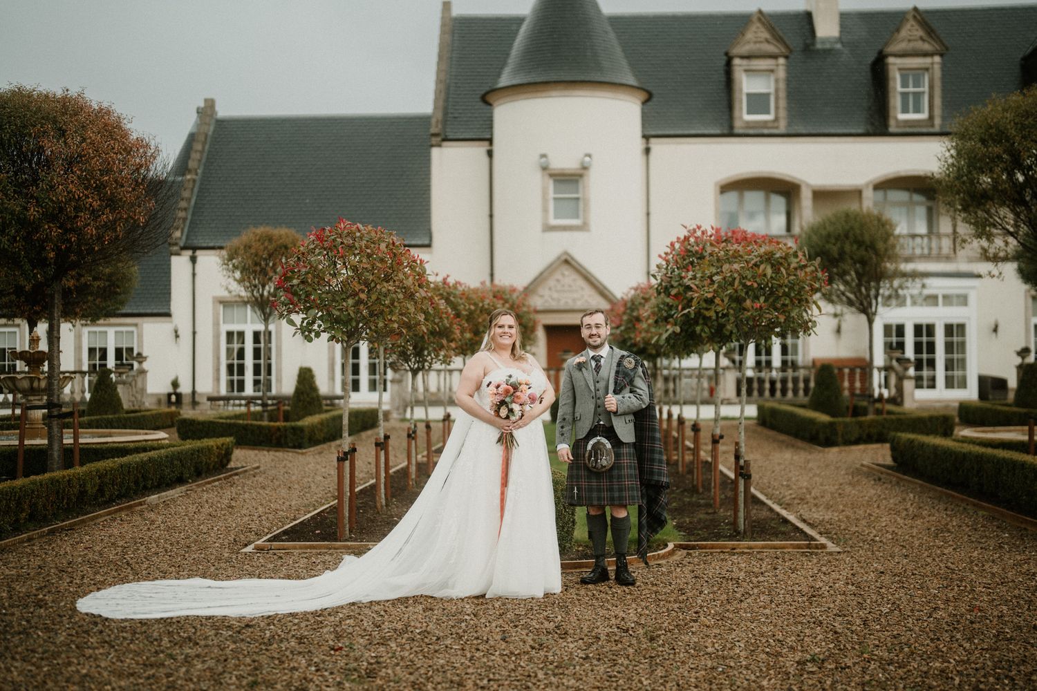 A Scottish wedding portrait in front of an elegant white manor house with formal gardens.
