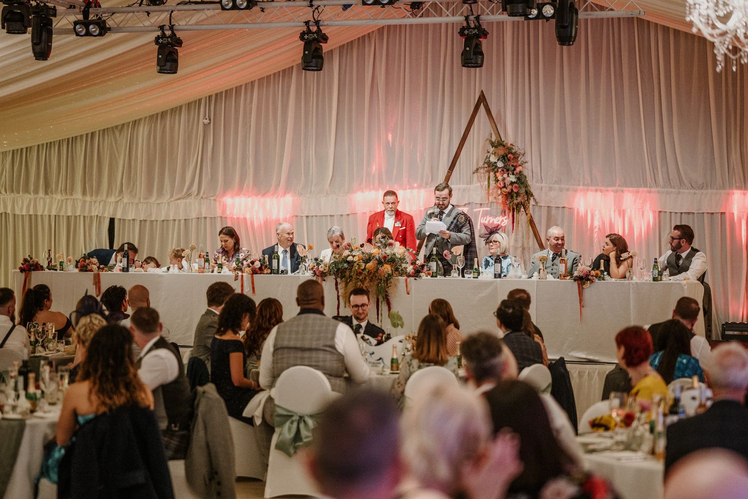 Wedding venue with elevated head table and pink uplighting on white draped walls.