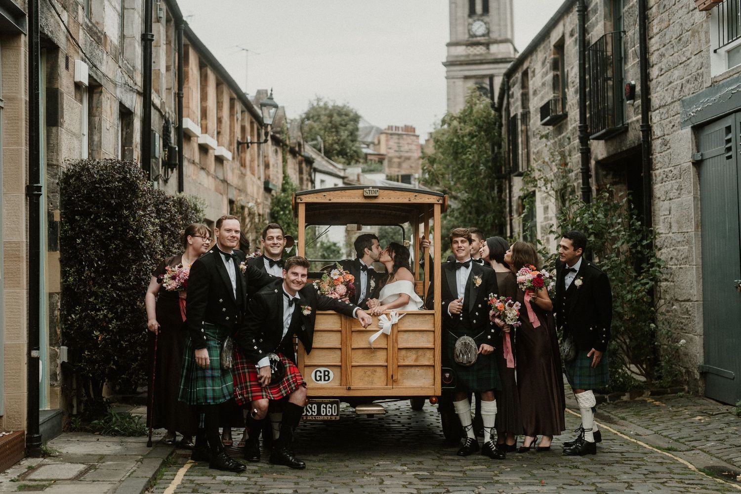 Wedding party surrounding wooden cart on cobblestone street with traditional Scottish kilts.