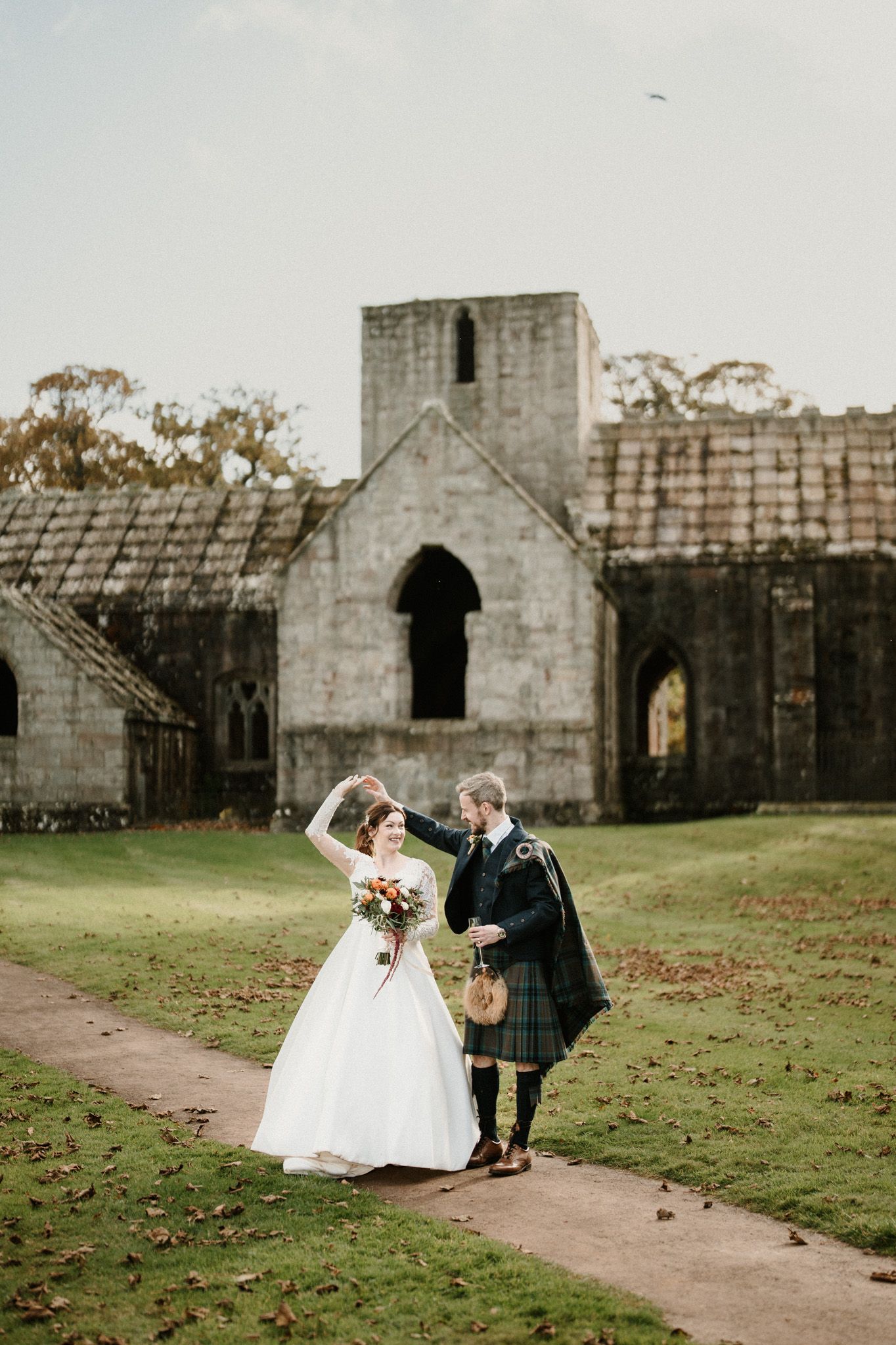 Wedding couple walks along path near historic stone ruins in Scottish countryside.