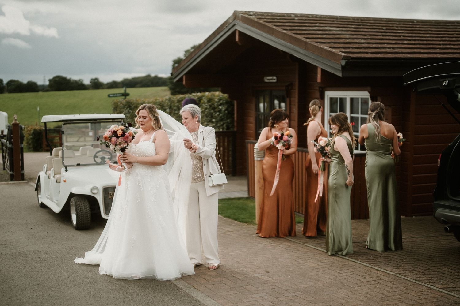 Wedding party members in coordinated dresses gather outside rustic venue.