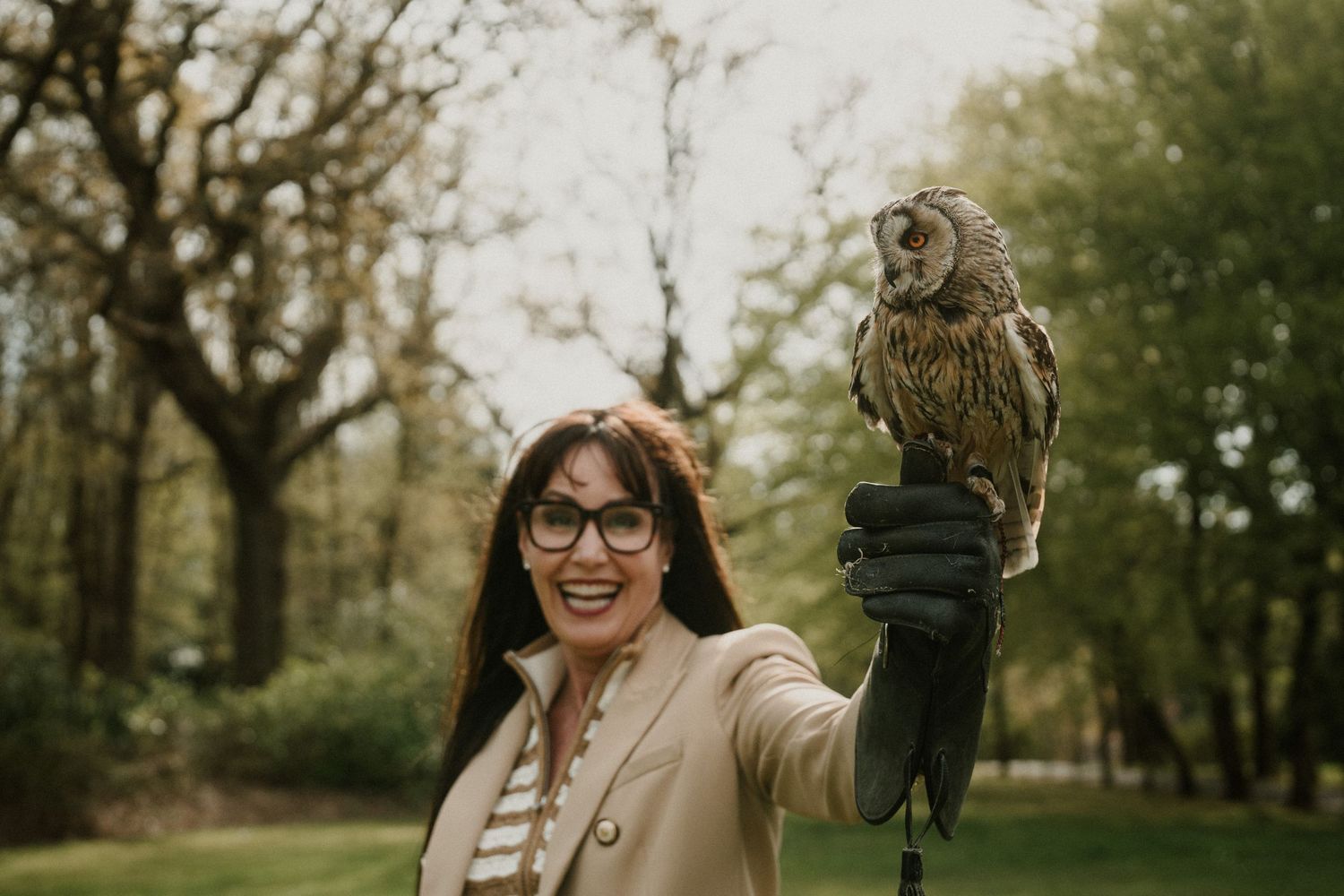 A person in a beige coat holds a tawny owl on their gloved hand in an outdoor park setting.