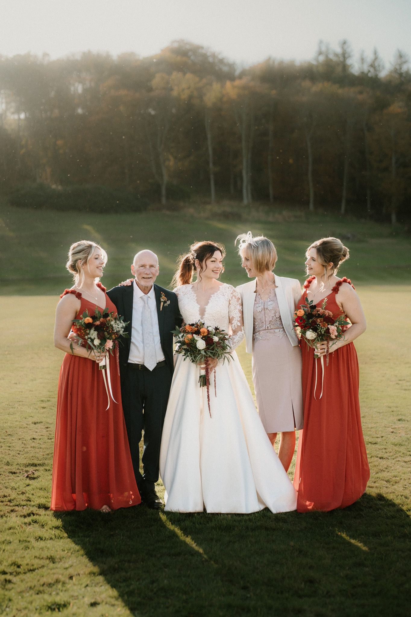 Wedding party in red and neutral formal attire gather for sunset portrait on grassy hillside.