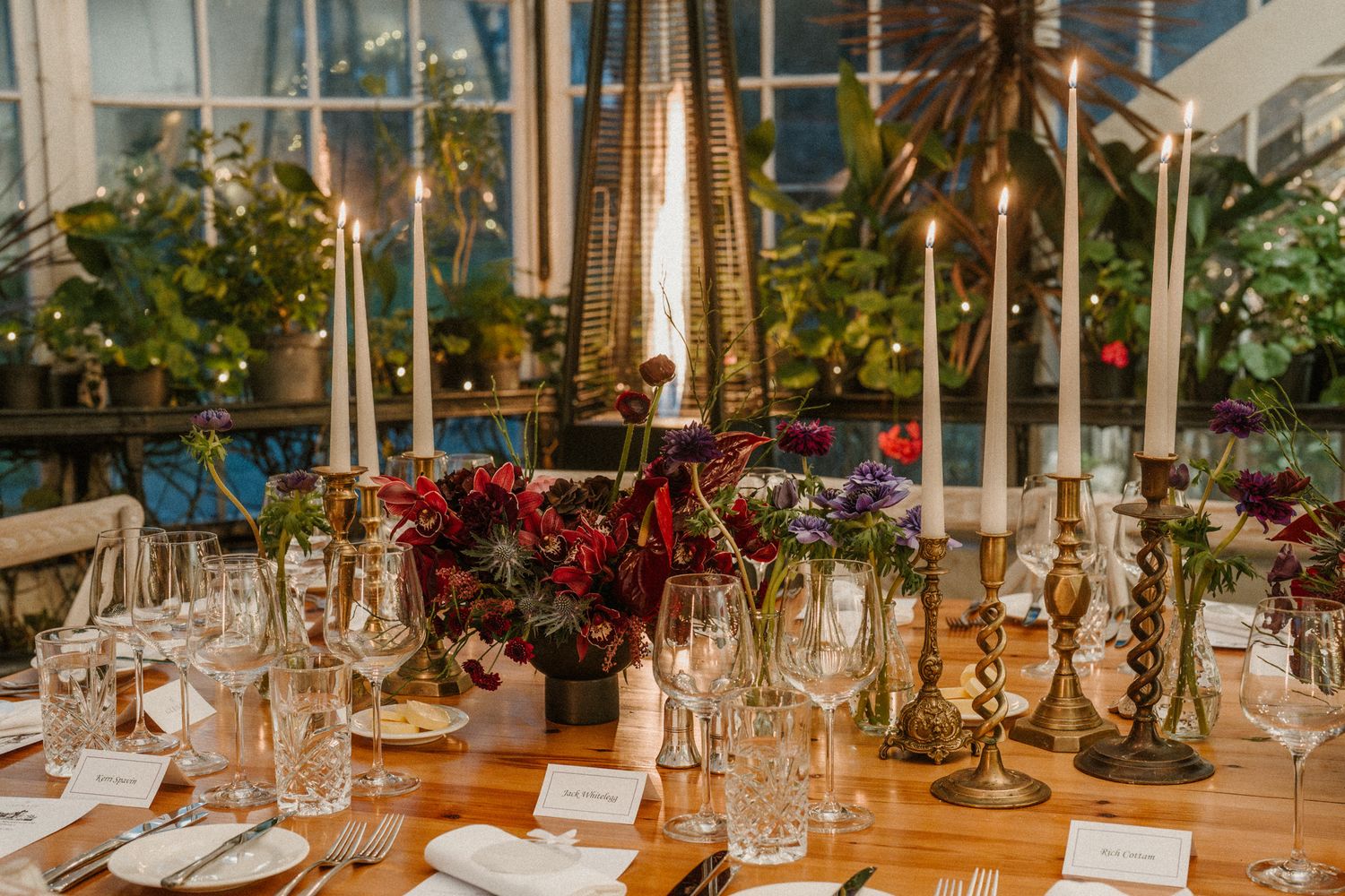 Romantic table decor featuring brass candlesticks, burgundy flowers, and place settings in a glass-enclosed dining space.