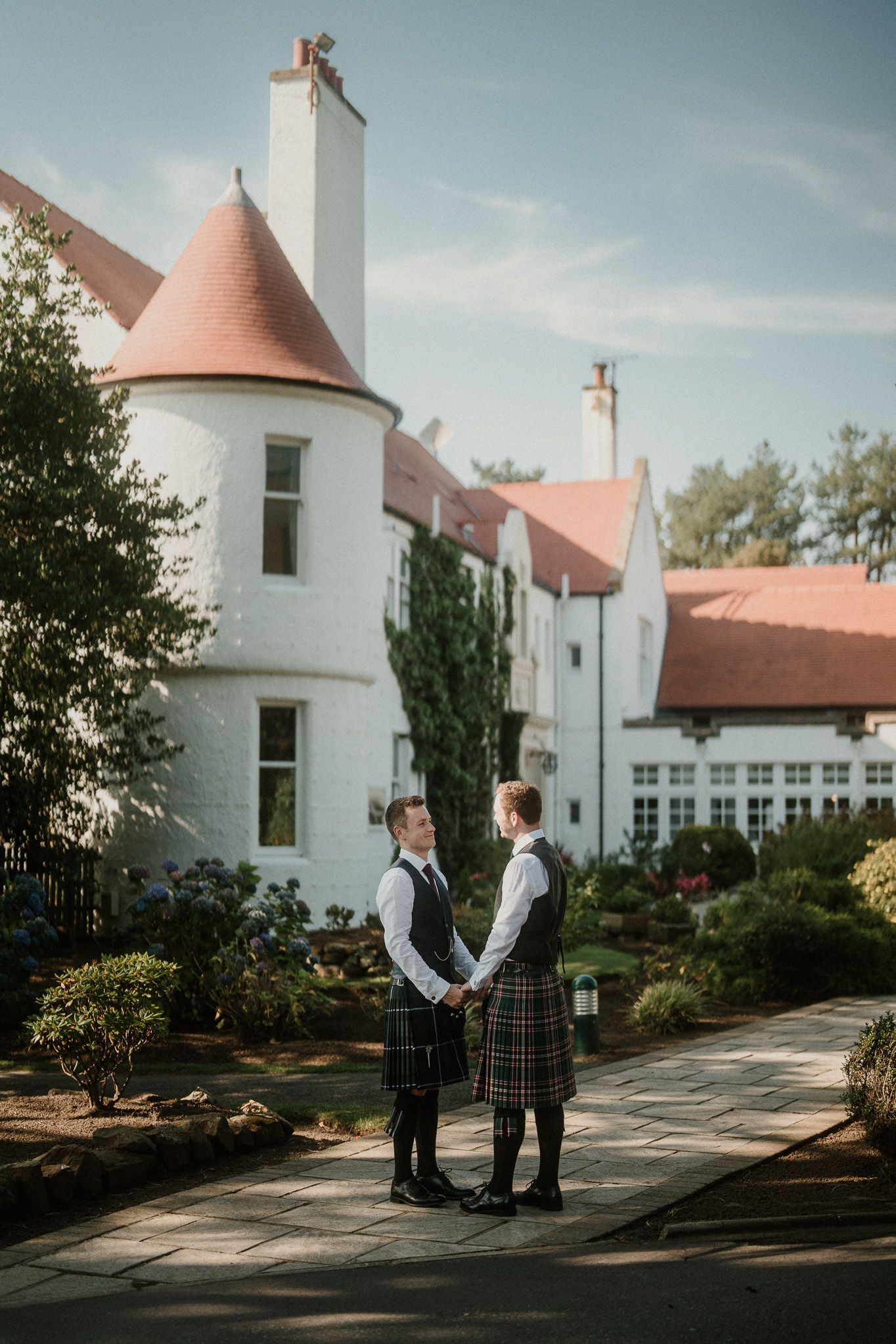 A couple in Scottish attire stands together outside a white castle-style building with terracotta roofs.