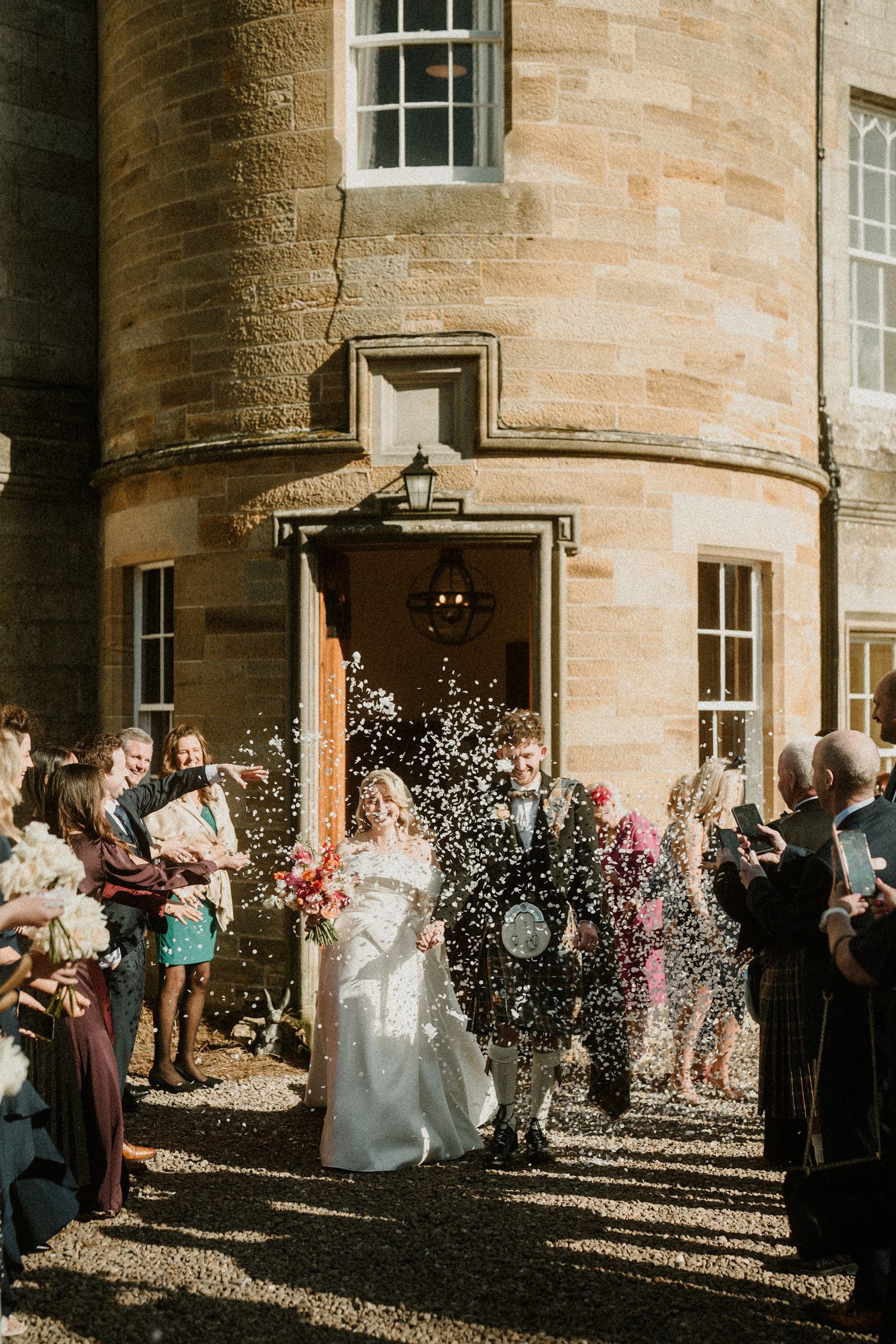 Guests throw confetti over newlyweds outside stone castle entrance in bright sunlight.