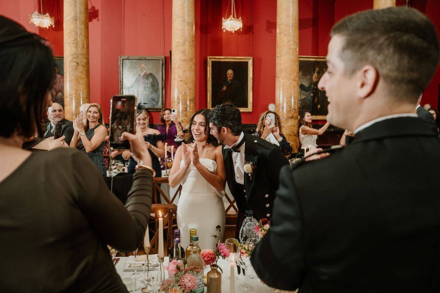 Wedding guests seated at long tables in a grand ballroom with red walls and portrait-lined walls.