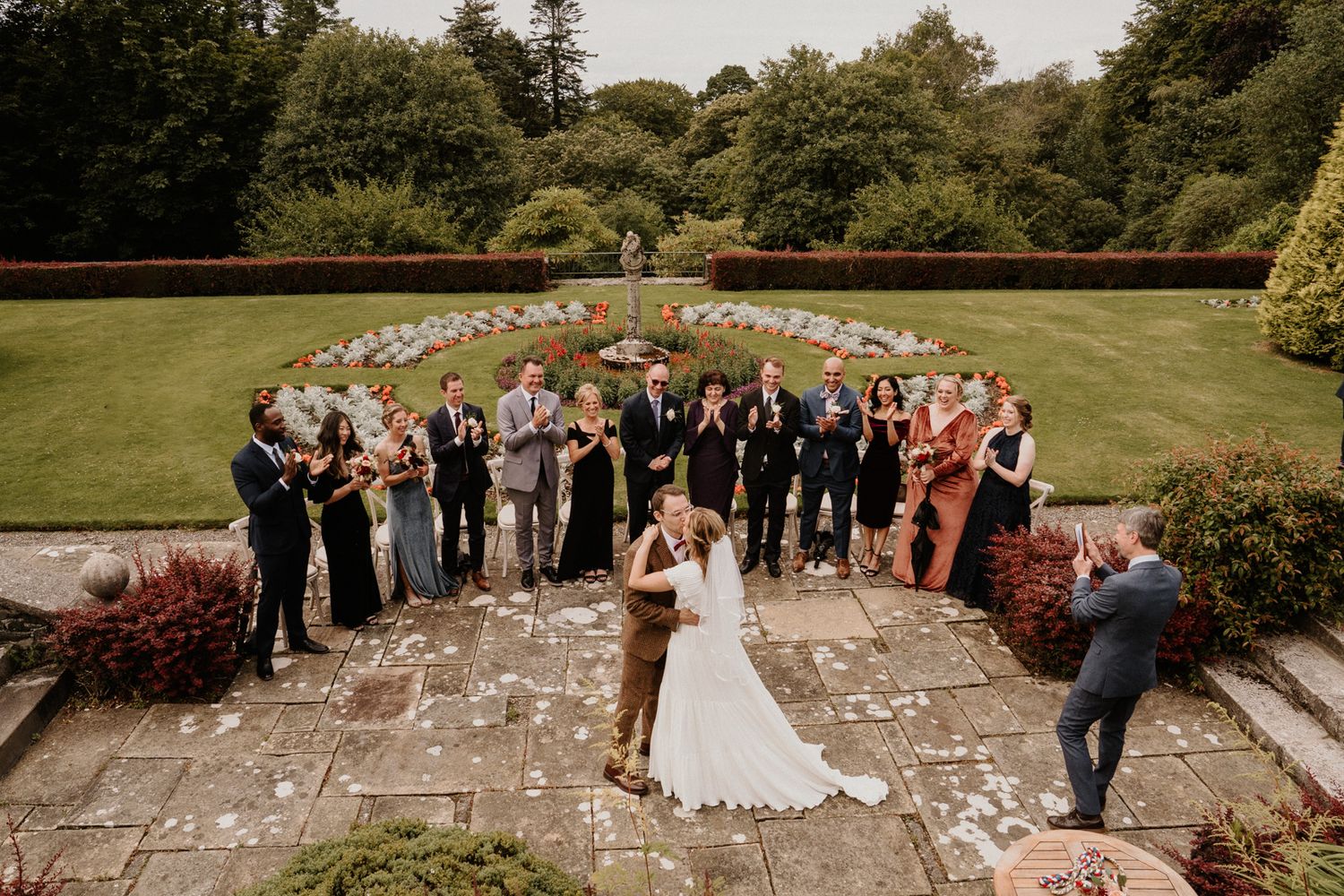 Wedding celebration with guests forming heart shape on stone patio in garden setting.