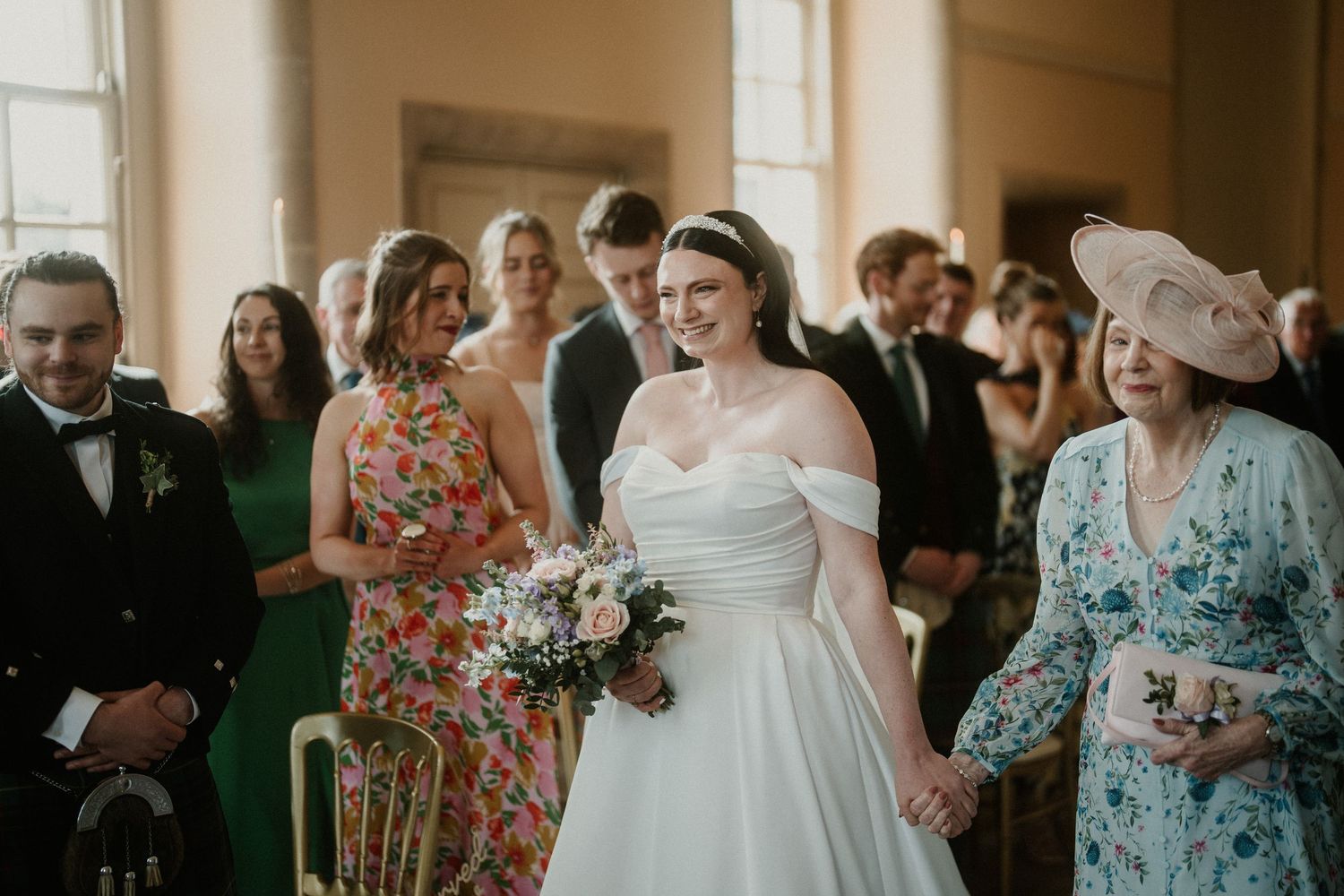Wedding party members in floral dresses and formal attire gather during an indoor celebration.
