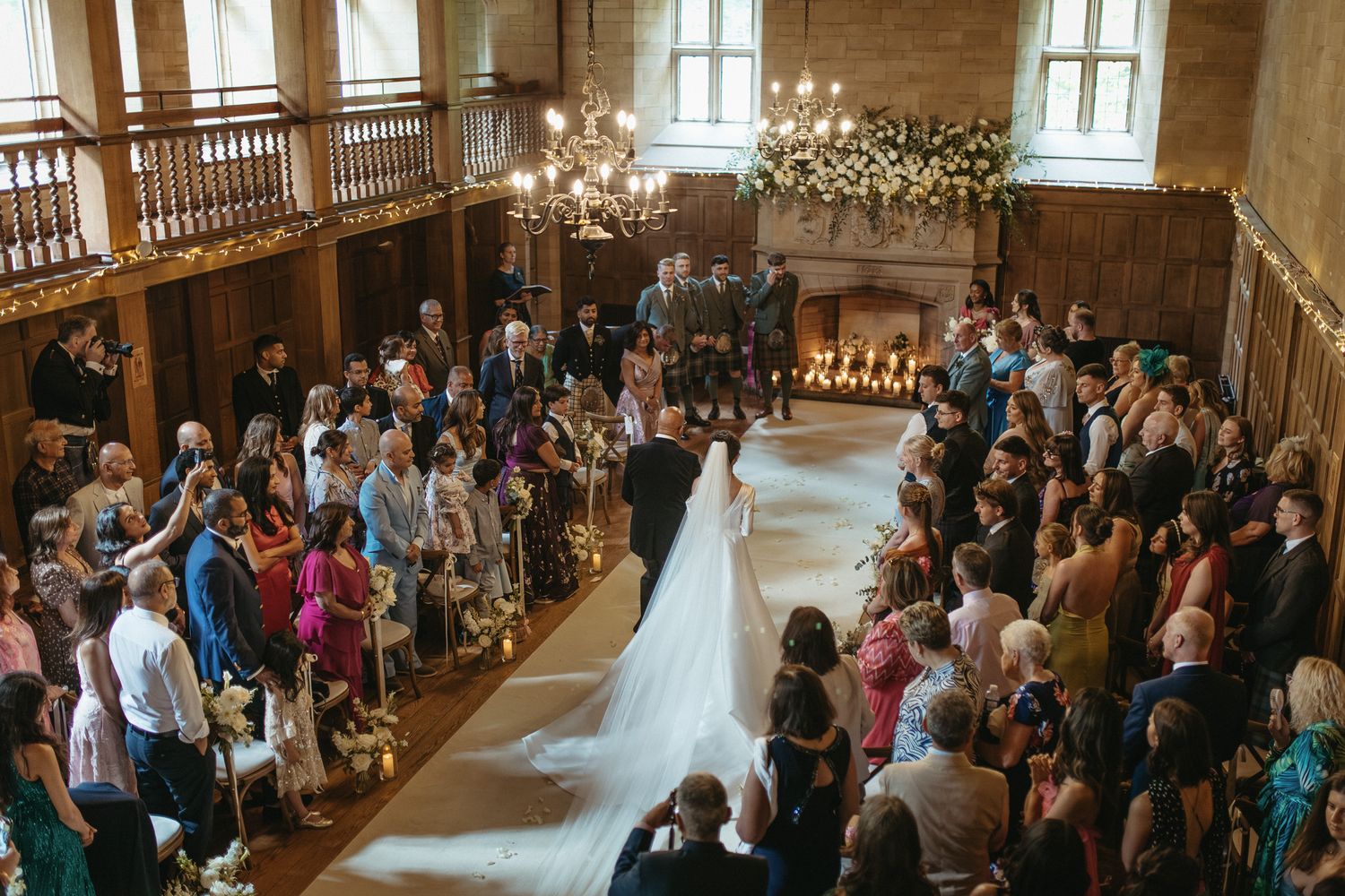 Elegant wedding ceremony in a grand hall with guests lining the aisle as bride walks toward floral altar.